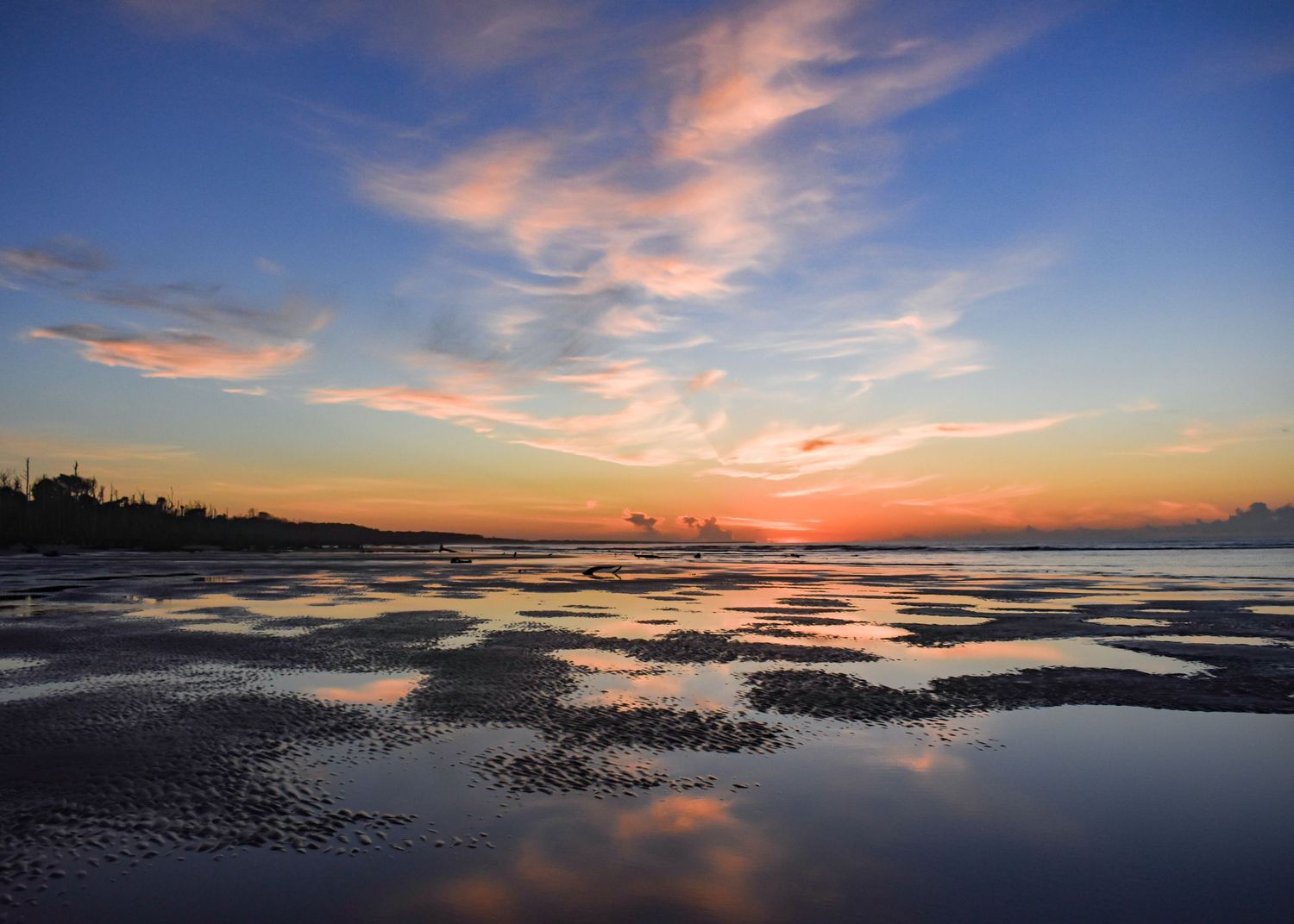 Colorful sunset reflects on wet tidal flats with pink and blue clouds stretching across the coastal horizon.