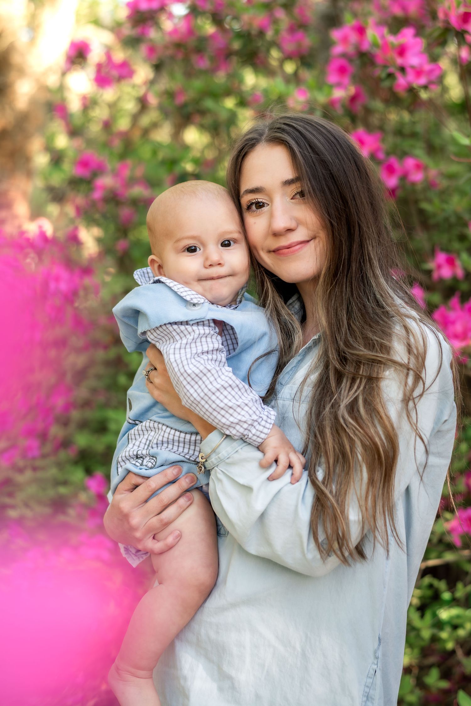 Young woman with long brown hair holds a smiling baby boy outdoors near vibrant pink flowering bushes.