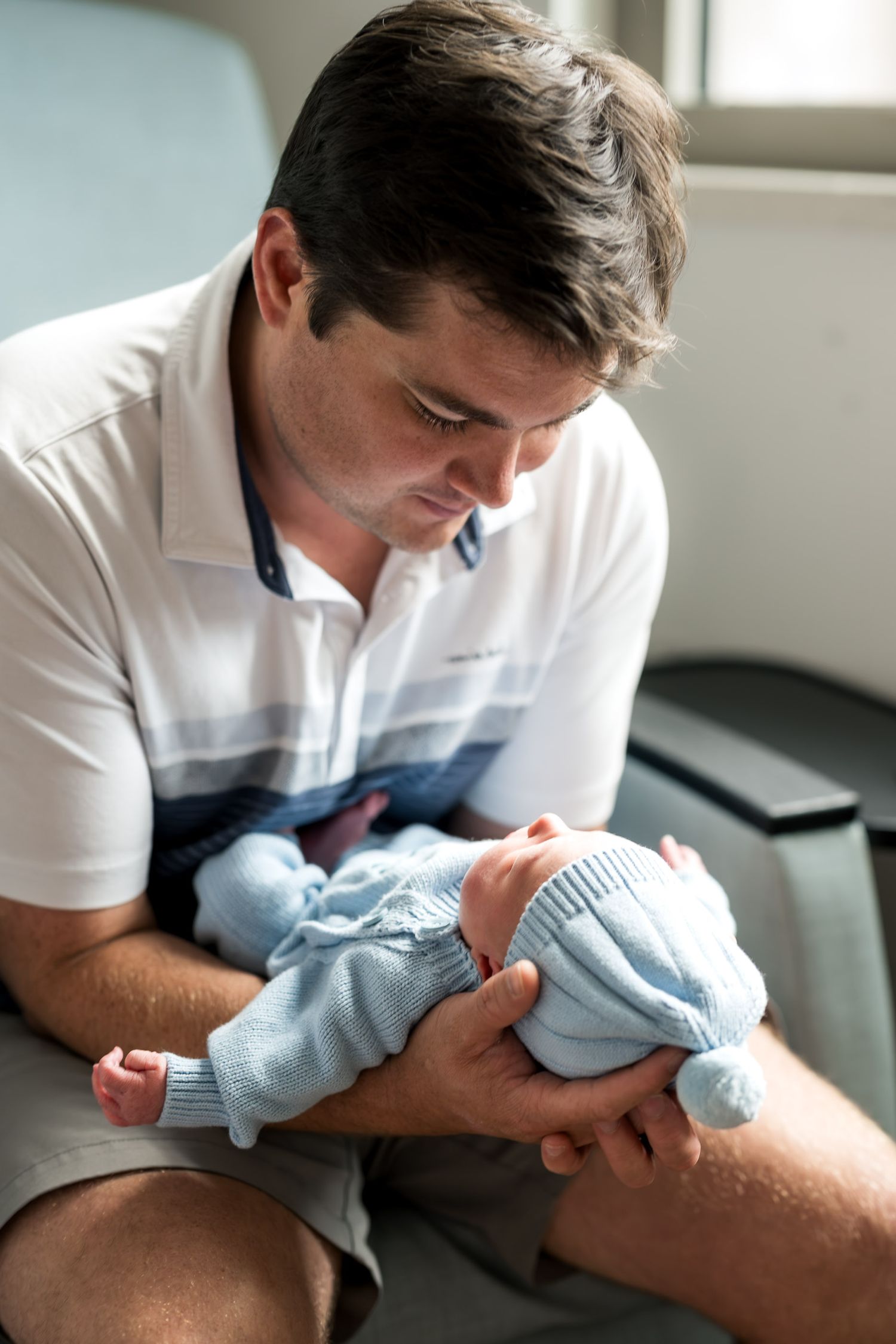 A father tenderly holds his newborn baby dressed in a light blue knit outfit while sitting indoors.