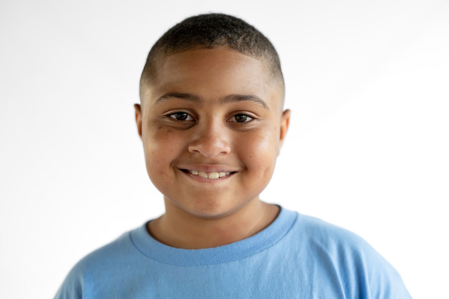 Smiling young boy with short hair wearing a light blue t-shirt against a white background.