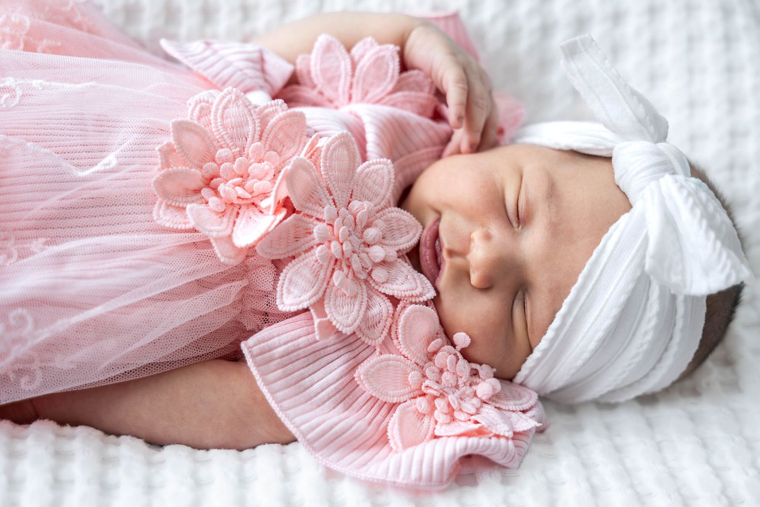 Sleeping newborn baby girl wearing a pink floral outfit and white bow headband on a white blanket.