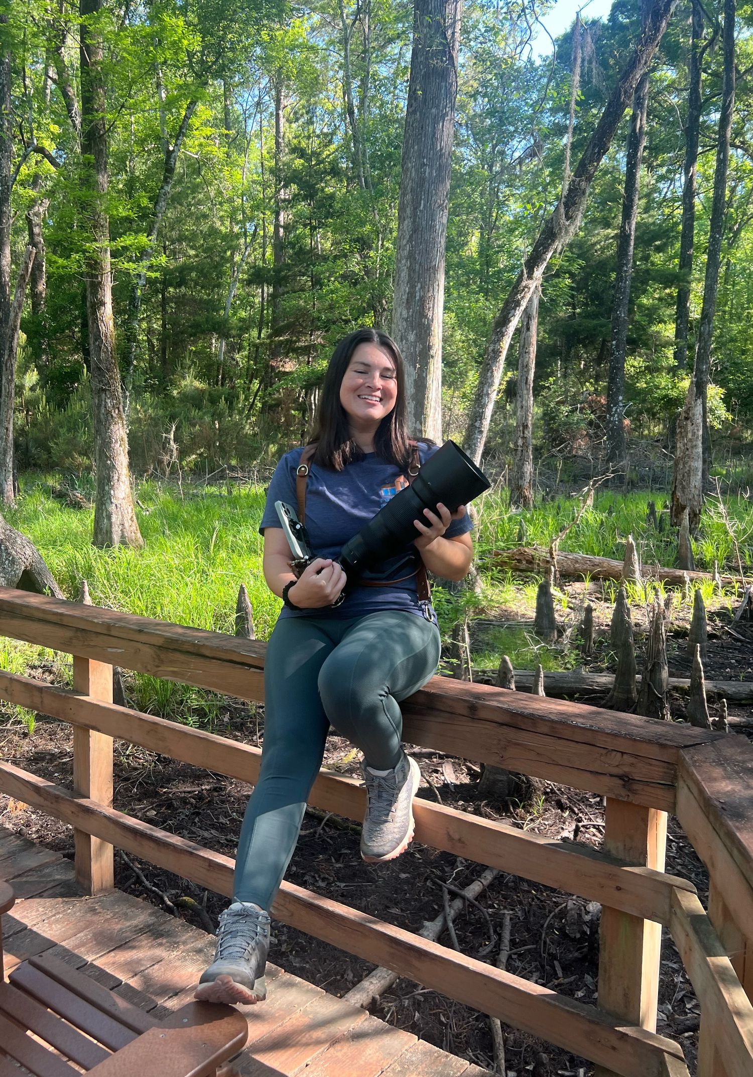 Young woman smiling on wooden boardwalk in lush green forest holding camera equipment outdoors.