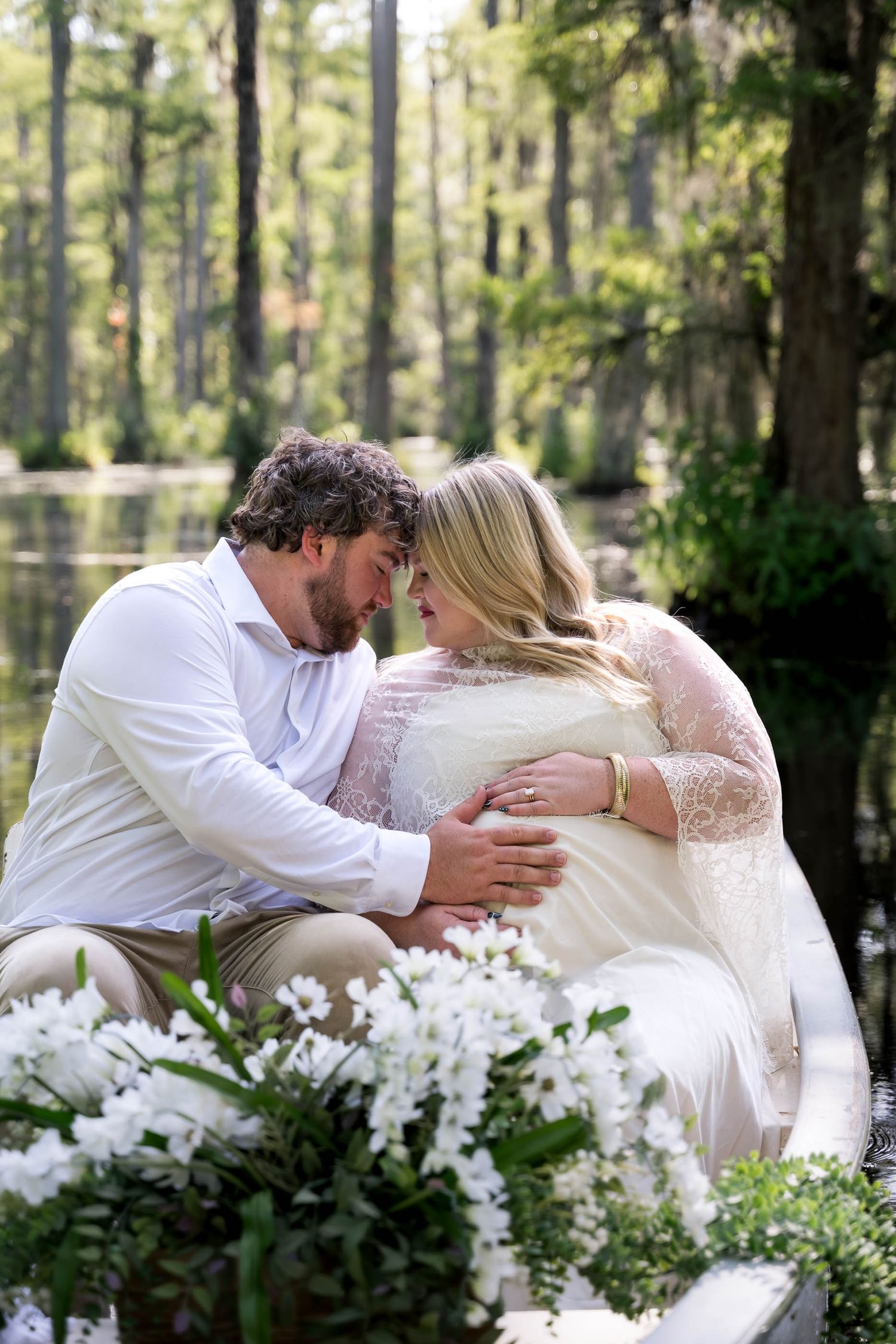 A couple in wedding attire share an intimate moment outdoors near a serene cypress swamp surrounded by white flowers.