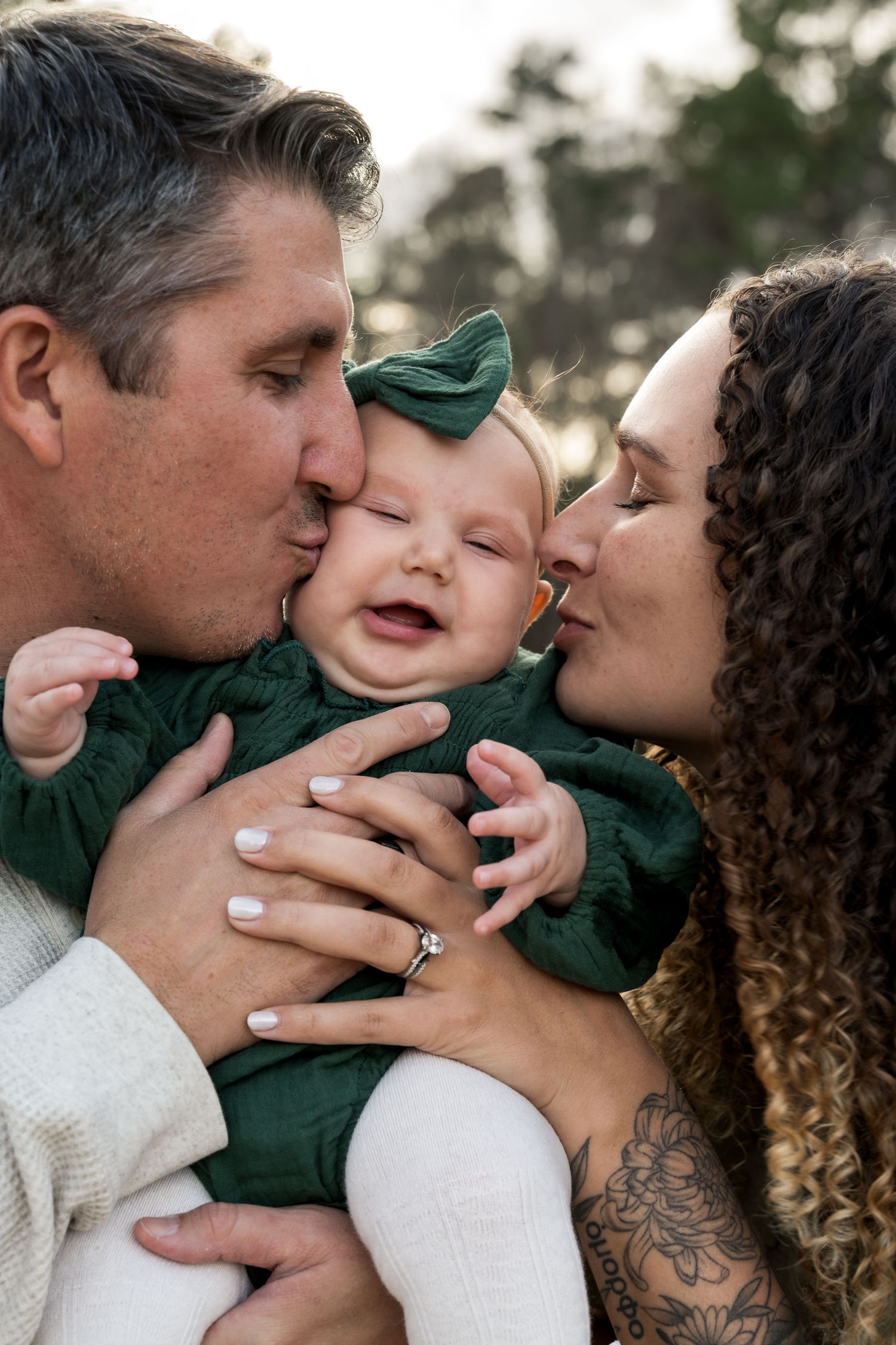 Parents kissing their smiling baby in a green outfit and bow during an outdoor family photo session.