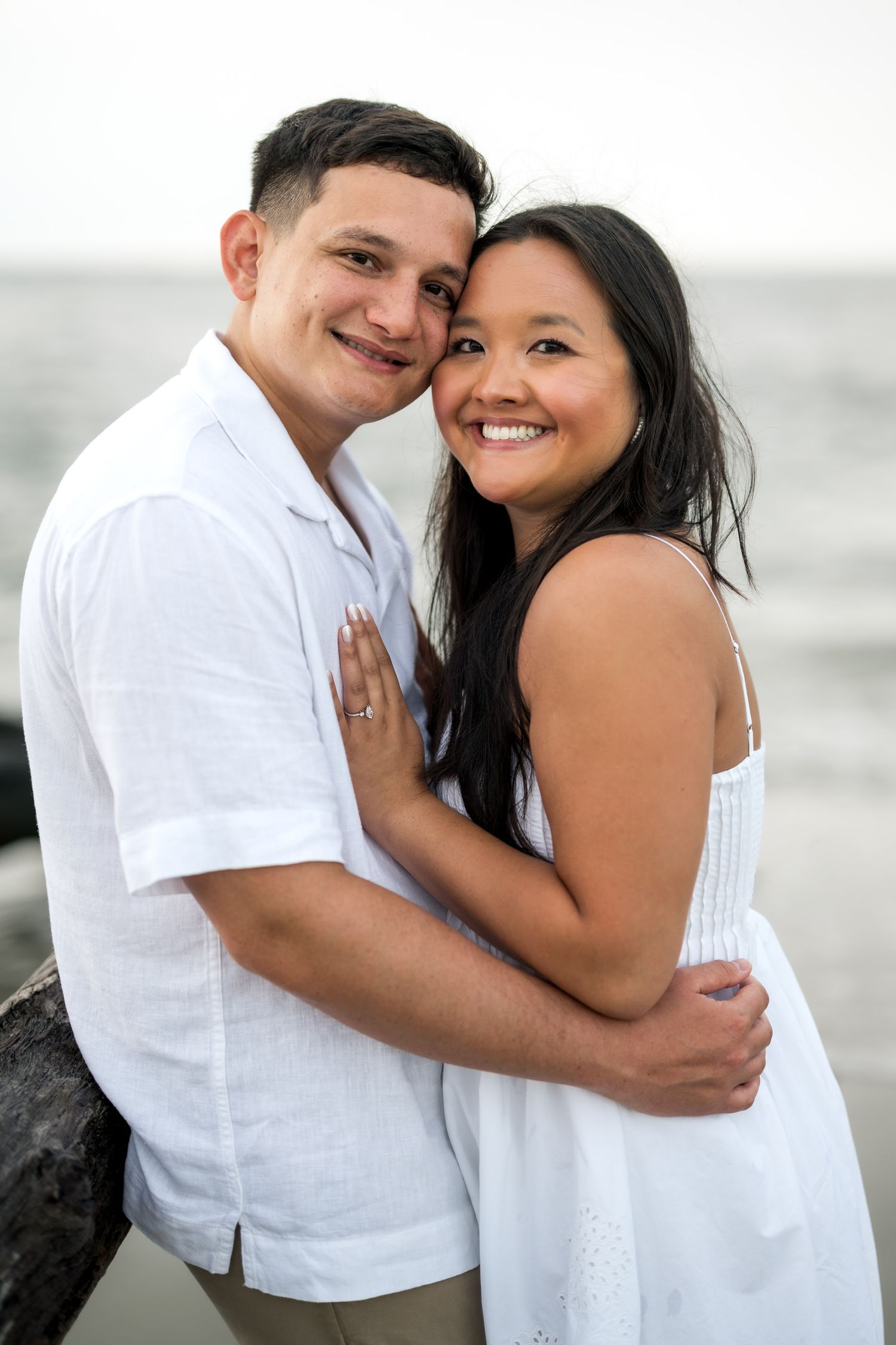 Smiling couple in matching white outfits embrace on a beach, woman showing engagement ring.