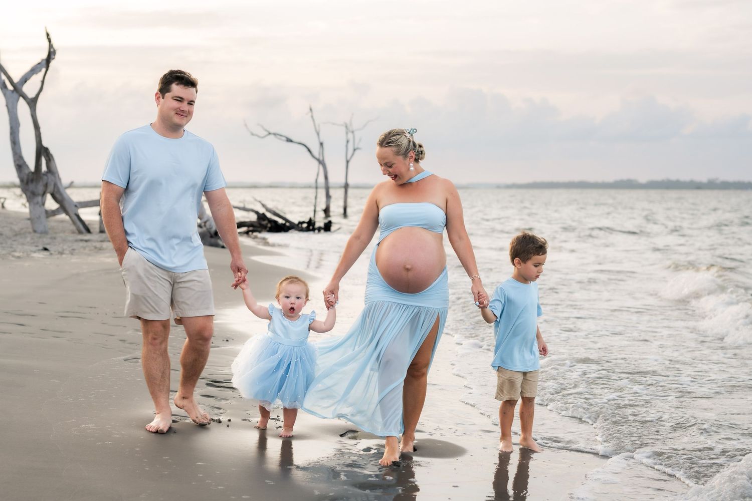 Pregnant family in blue outfits walking on beach, parents holding toddler's hands with young boy beside them.