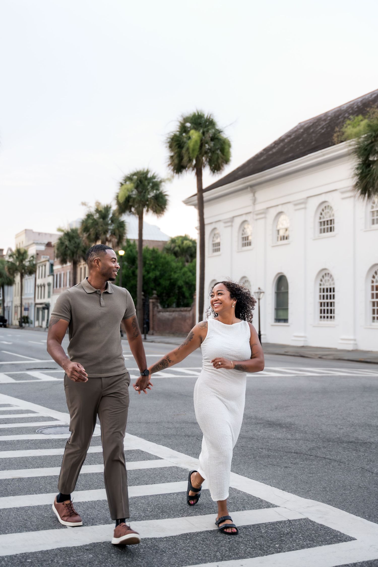 A joyful couple holds hands while crossing a street in Charleston, SC, with palm trees and a white building behind them.