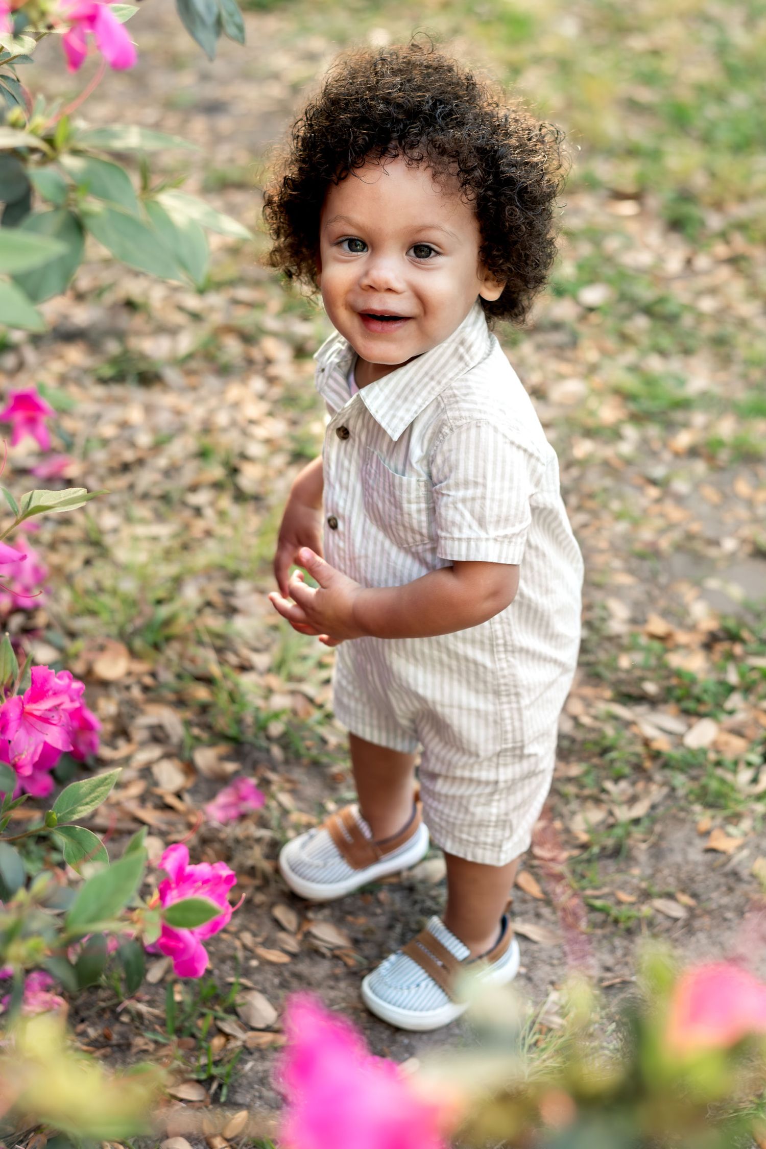 Hampton Park. Adorable curly-haired toddler boy smiling in striped outfit among pink flowers.