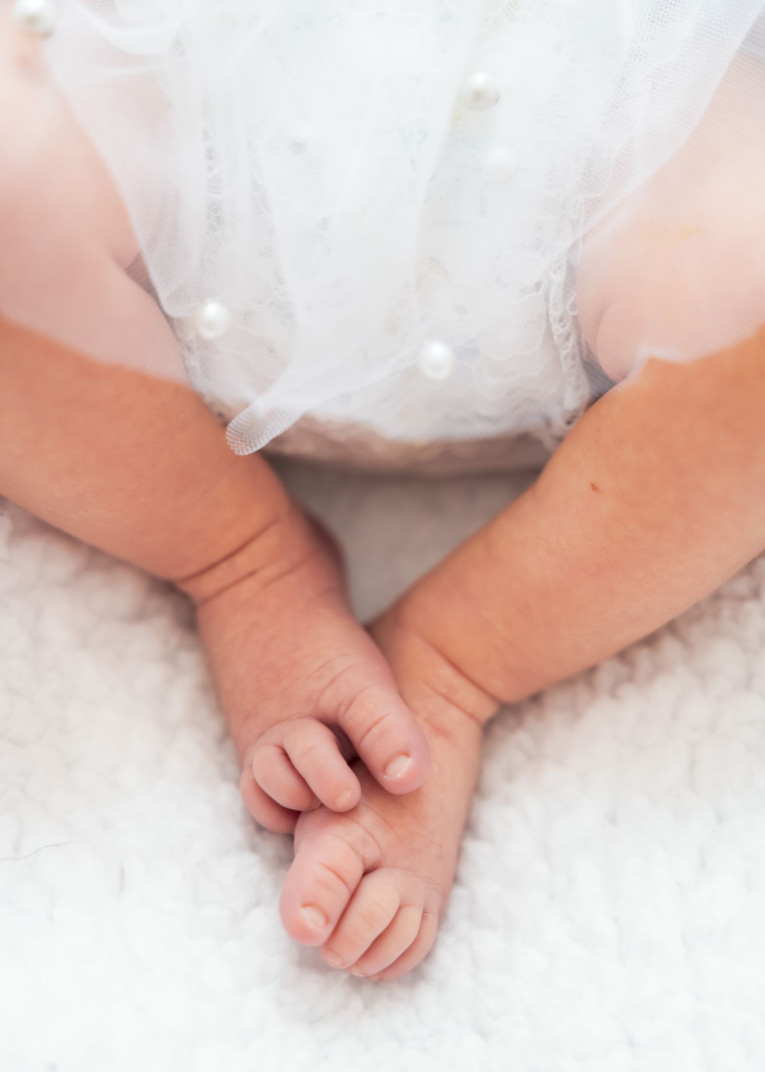 Close-up of a newborn baby's tiny feet and hands in a delicate white pearl-adorned outfit on soft fabric.