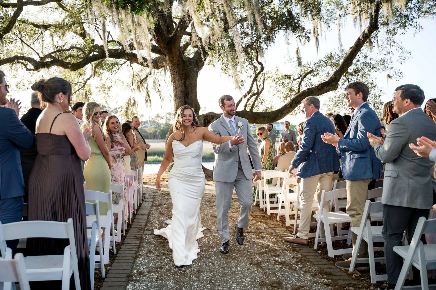 Dunes West Golf and RNewlyweds joyfully walking down outdoor aisle lined with white chairs and cheering guests under a moss-draped oak tree.