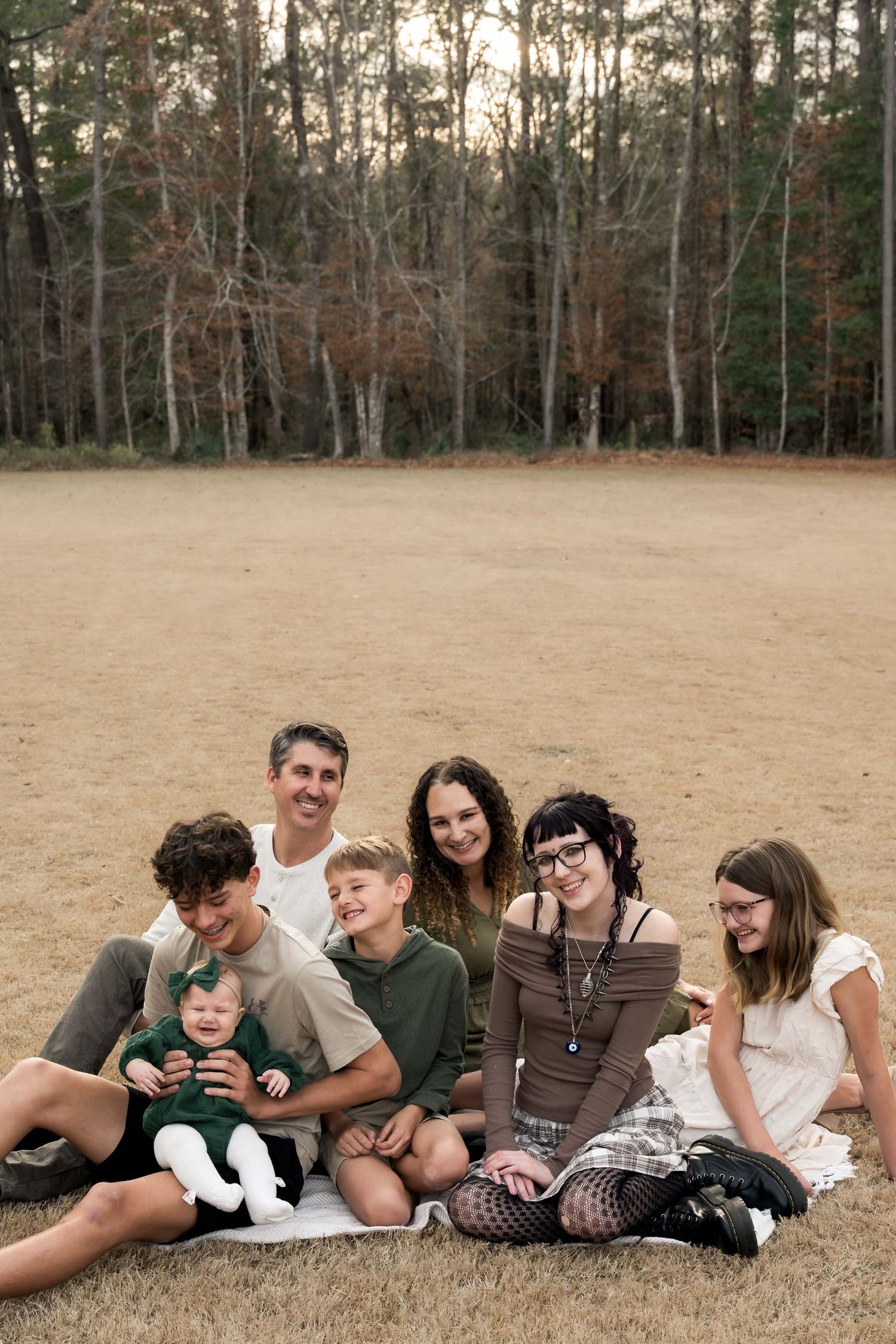 Happy family of seven sitting together on dry grass field with tall pine trees in background.