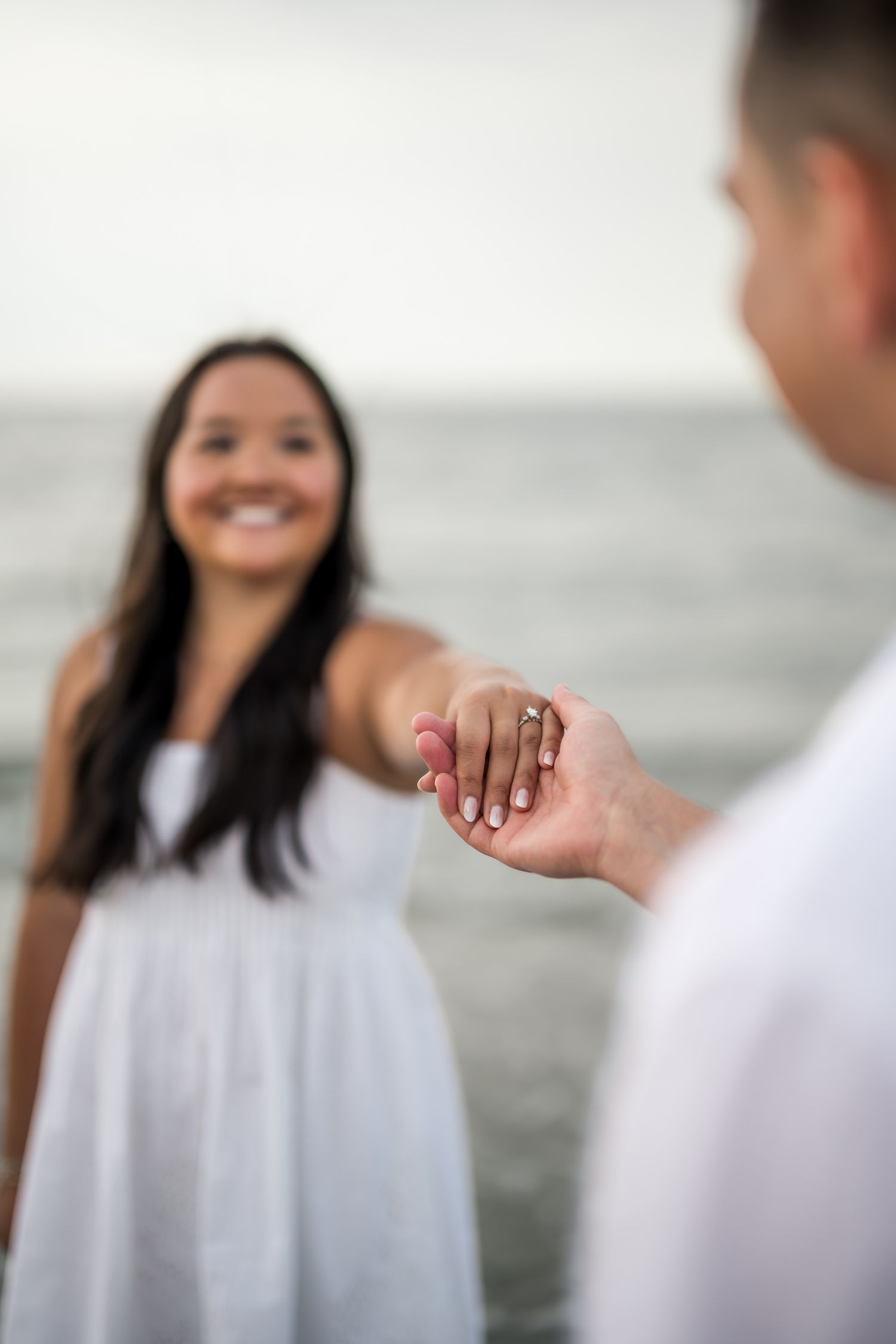 Close-up of couple holding hands showing engagement ring on beach with smiling woman in white dress.