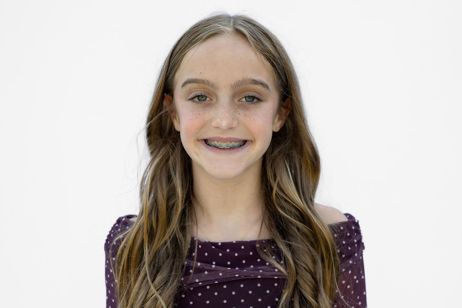 Smiling young girl with long brown hair and braces wearing a purple polka dot top against white background.