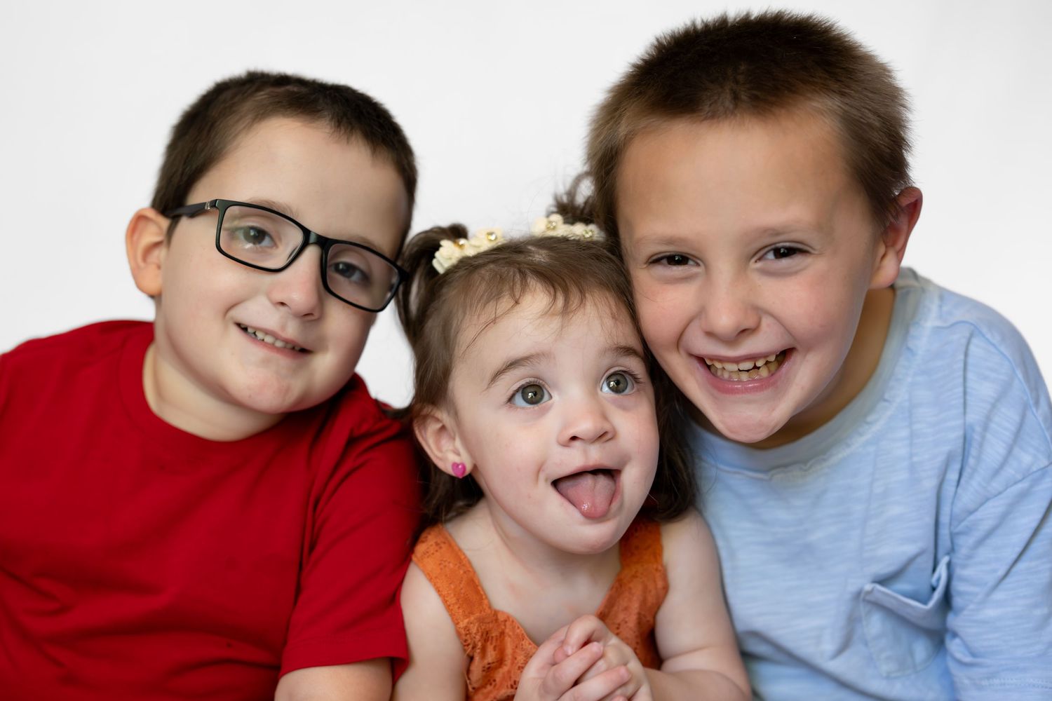 Three happy children pose together, two boys and a toddler girl sticking out her tongue, one boy wearing glasses.