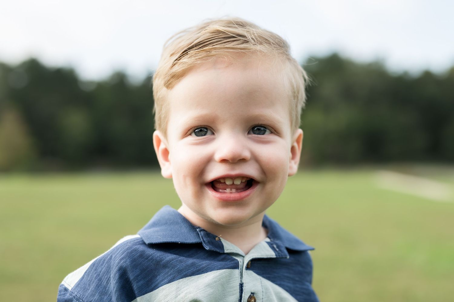 Smiling toddler boy with blonde hair wearing a blue jacket outdoors in a green field.