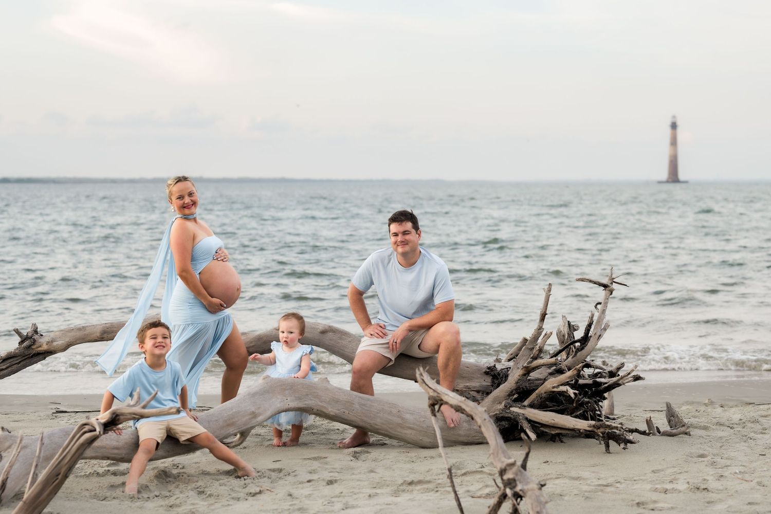 Pregnant family posing on driftwood at beach with lighthouse in background on a cloudy day.