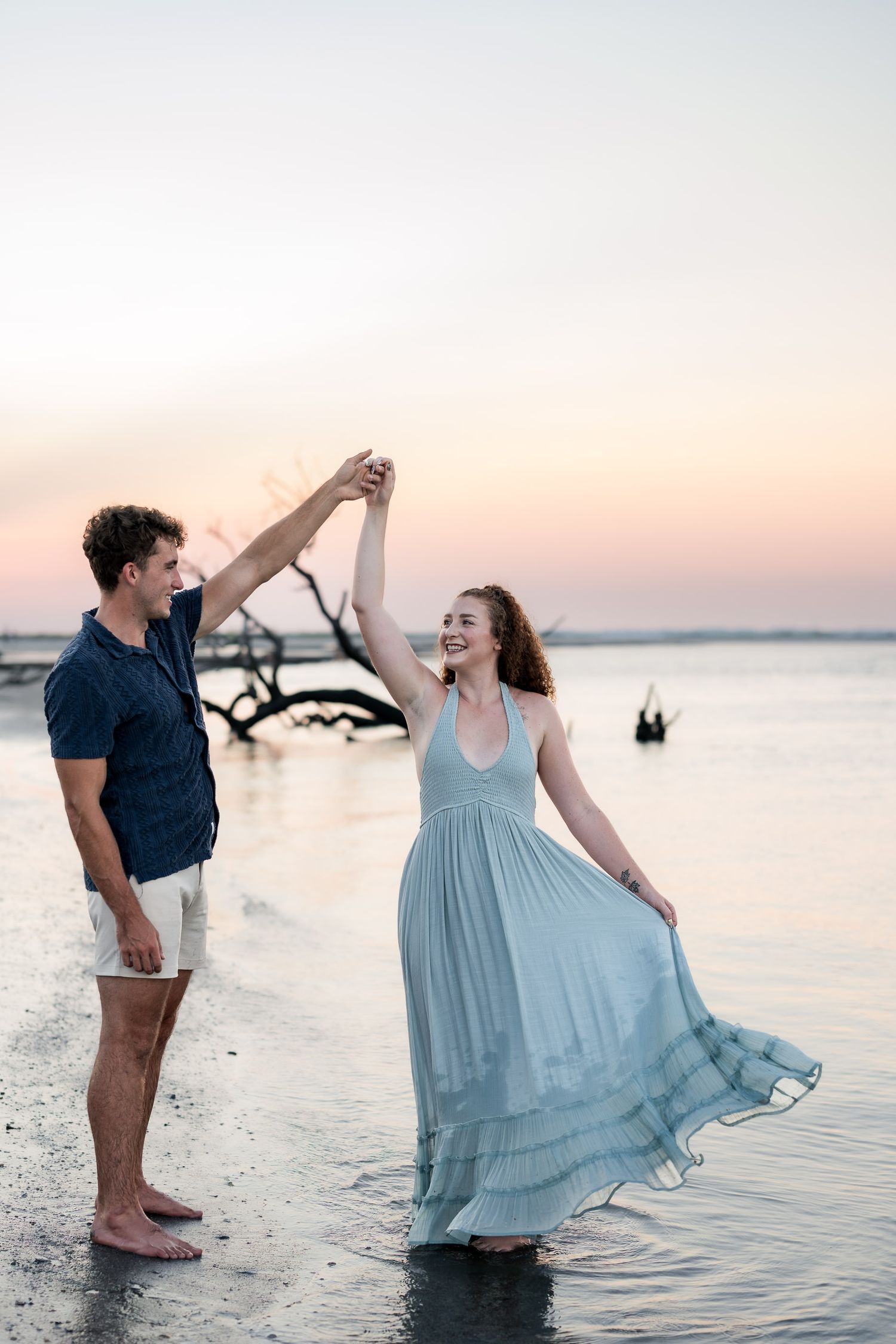 Couple dancing barefoot on a beach at sunset, man spinning woman in a flowing light blue dress near the water.