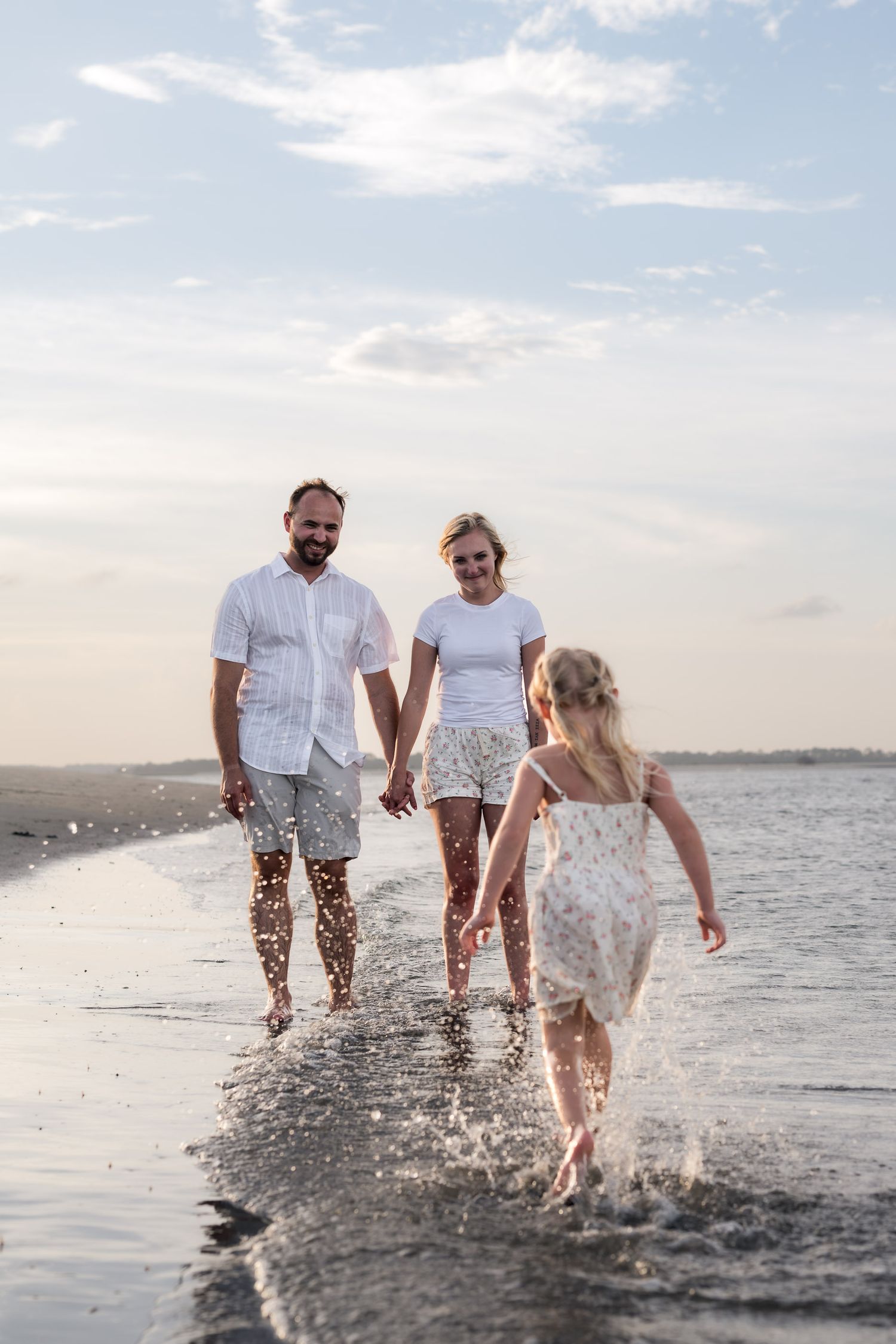 Family of three playing in ocean waves on a sandy beach at sunset, parents holding hands with young daughter splashing.