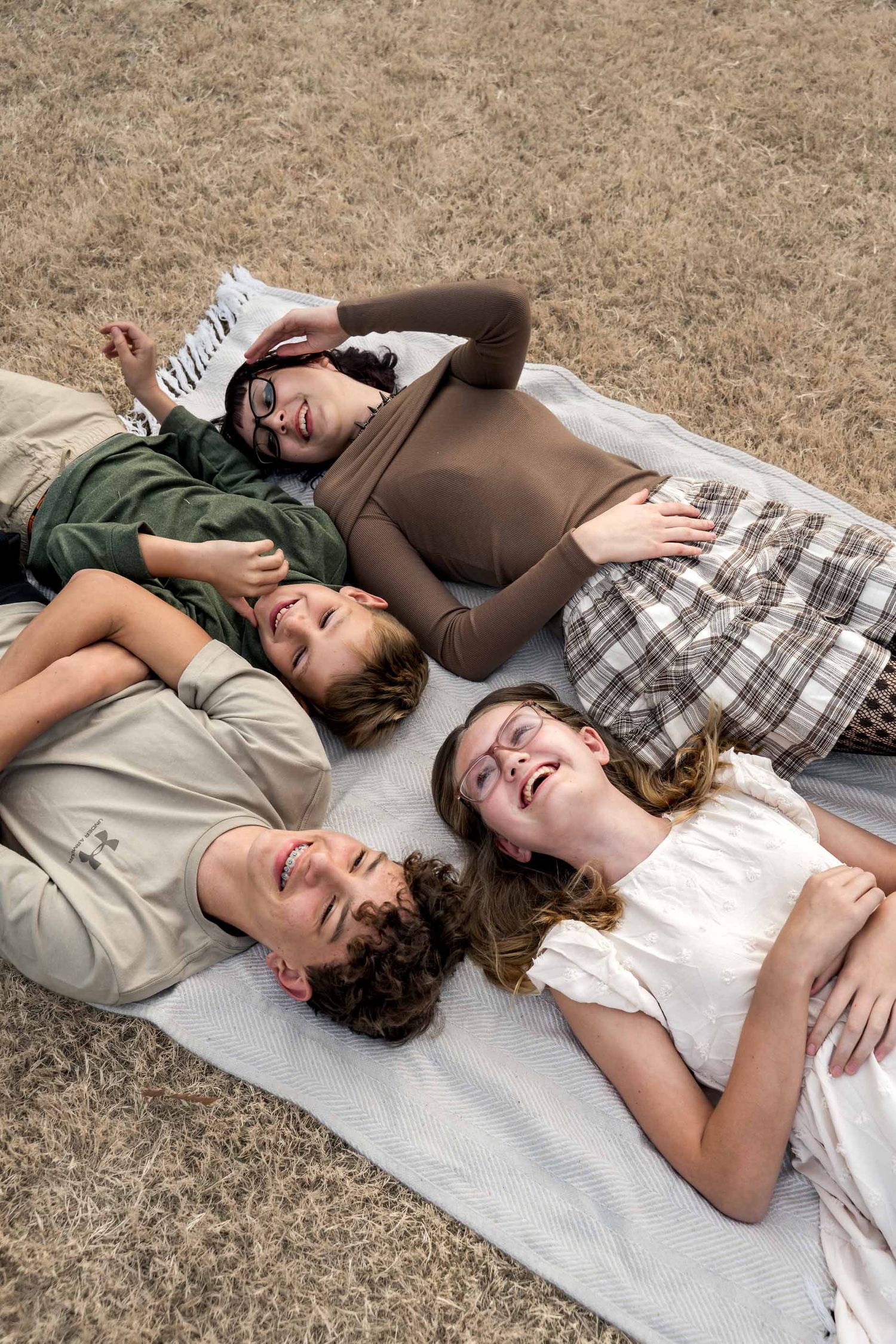 Four young people lying on a blanket outdoors, laughing and relaxing together on dry grass.