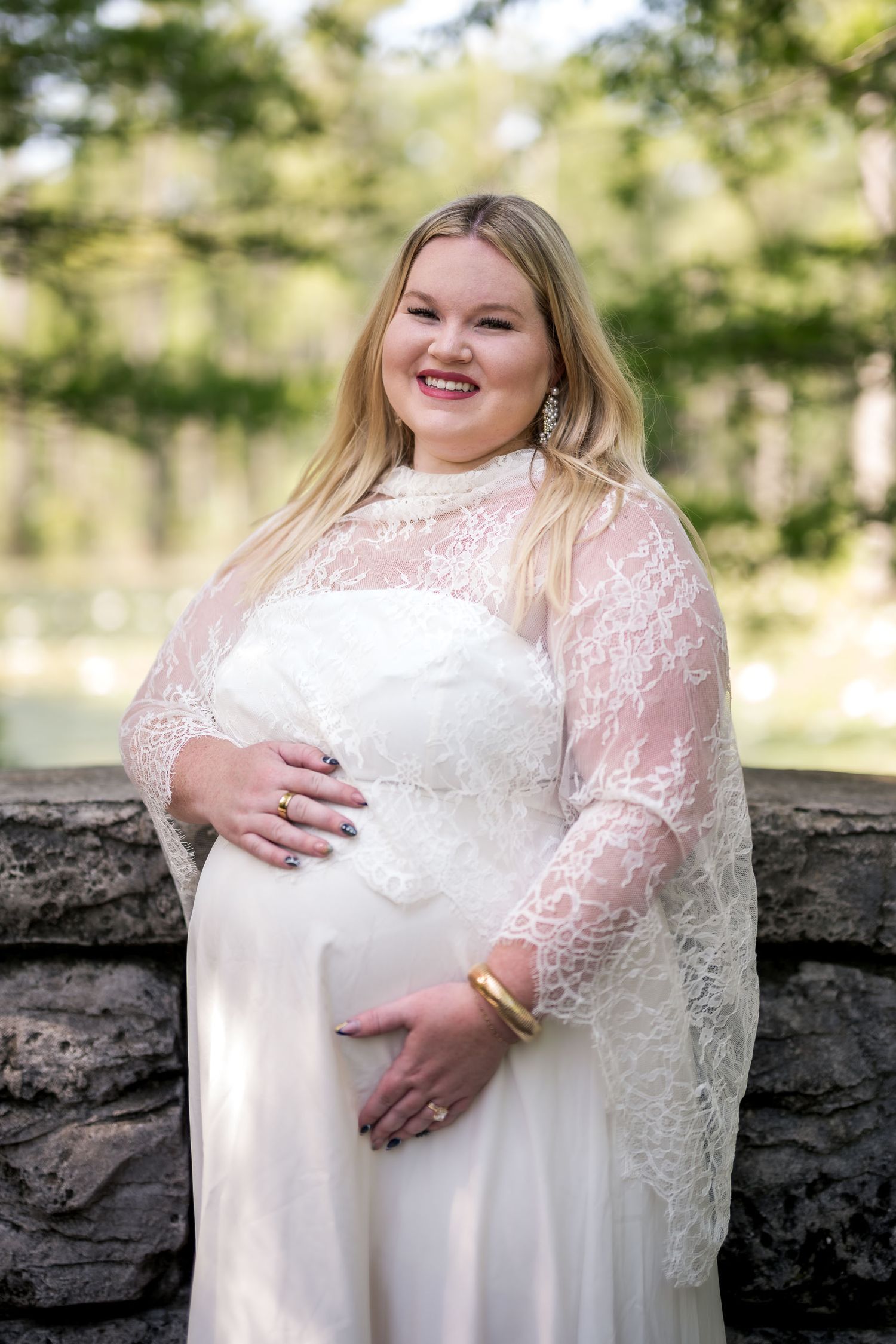 Pregnant bride in white lace long-sleeve wedding gown smiling outdoors near stone wall and trees.