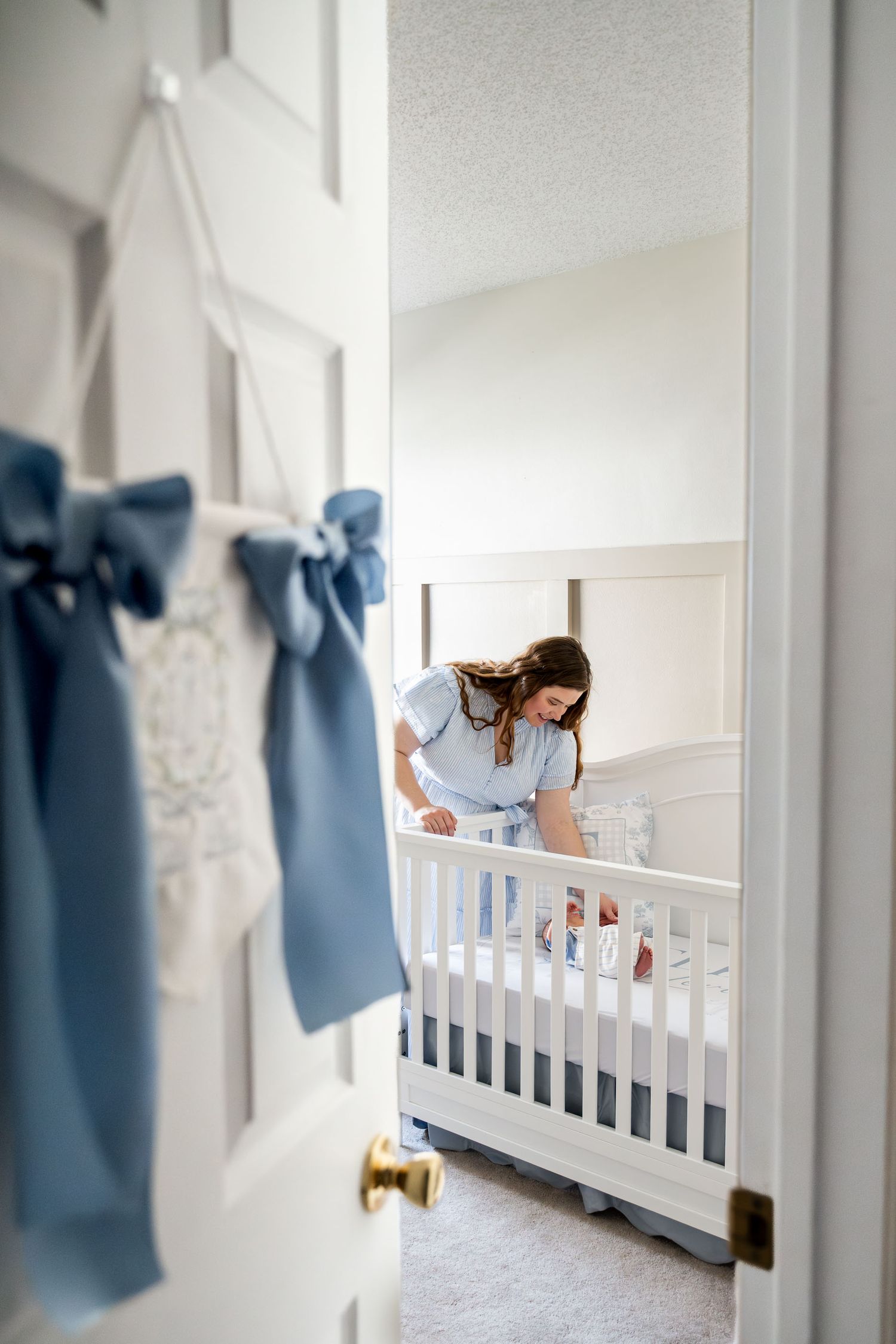 Mother leaning over white crib tending to newborn in soft blue nursery room.