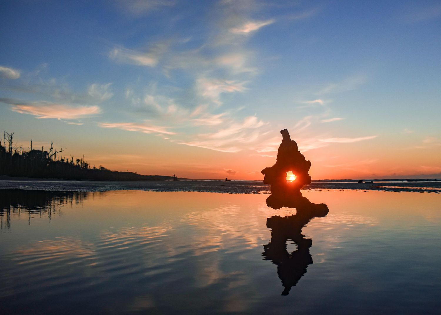 Silhouette of a person holding a camera at sunset on a reflective beach with colorful sky.