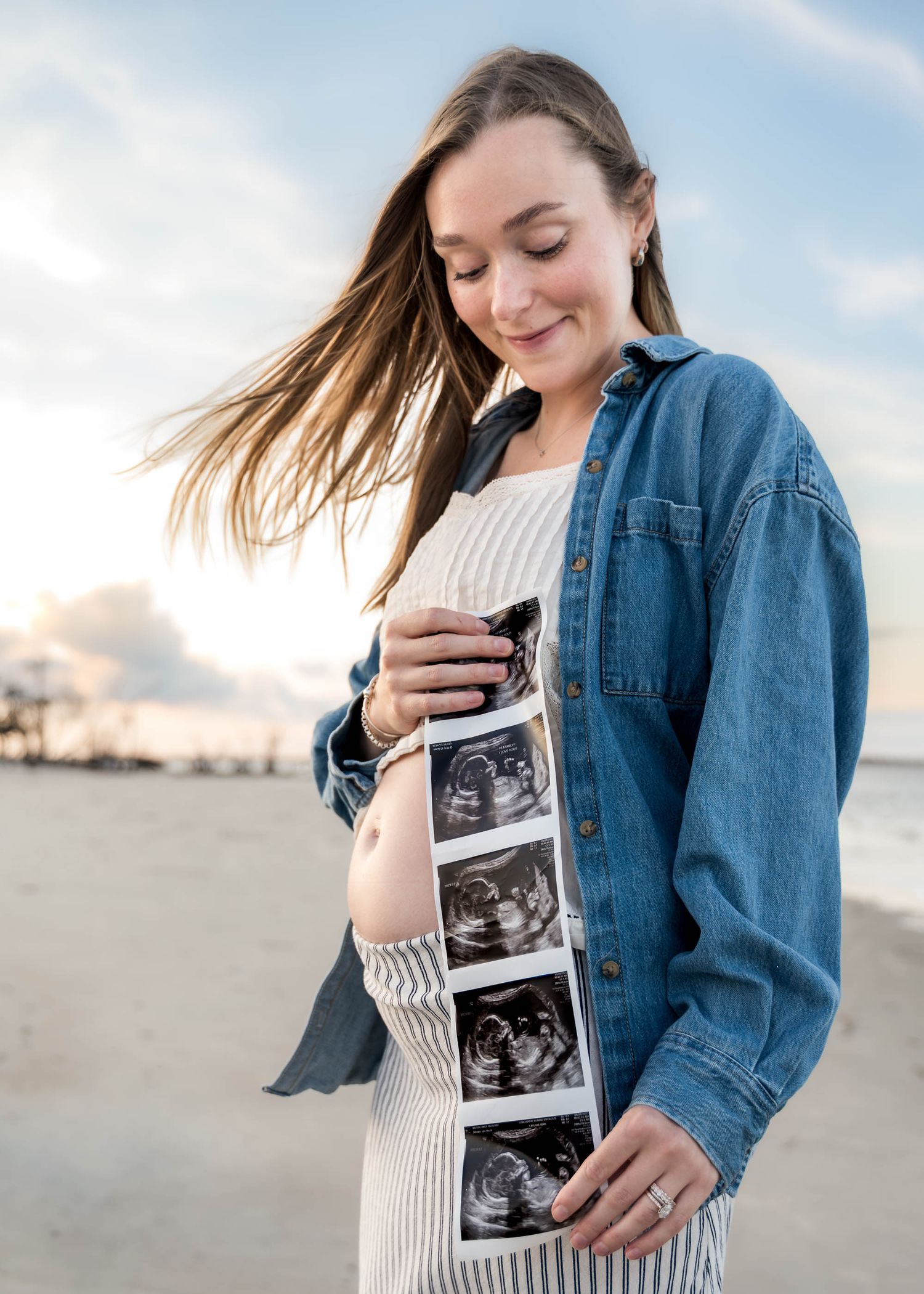 Folly Beach. Pregnant woman in denim jacket holding ultrasound strip photos on a breezy beach.
