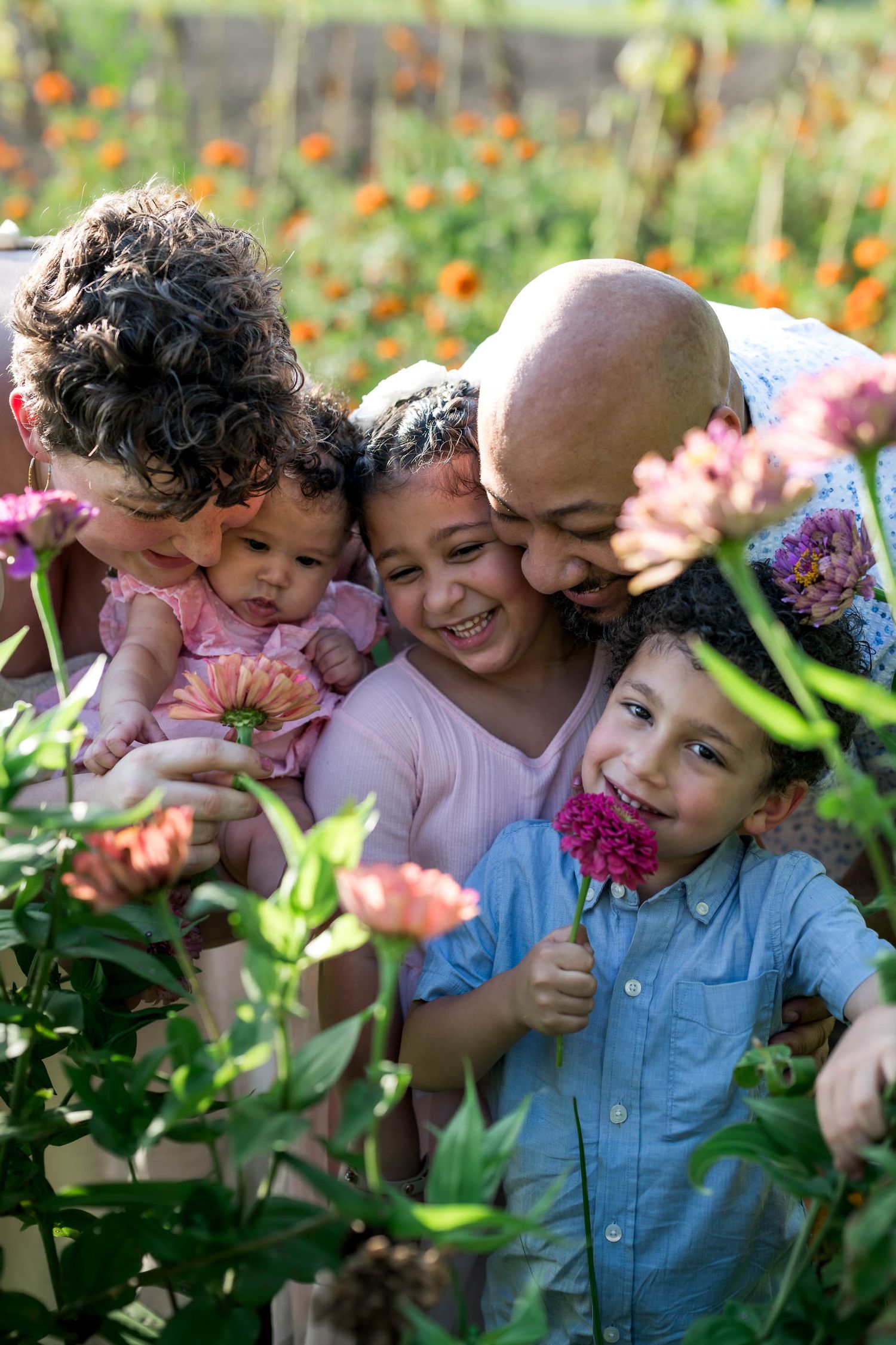 A joyful family with three young children smiles together among colorful blooming flowers in a sunny garden.