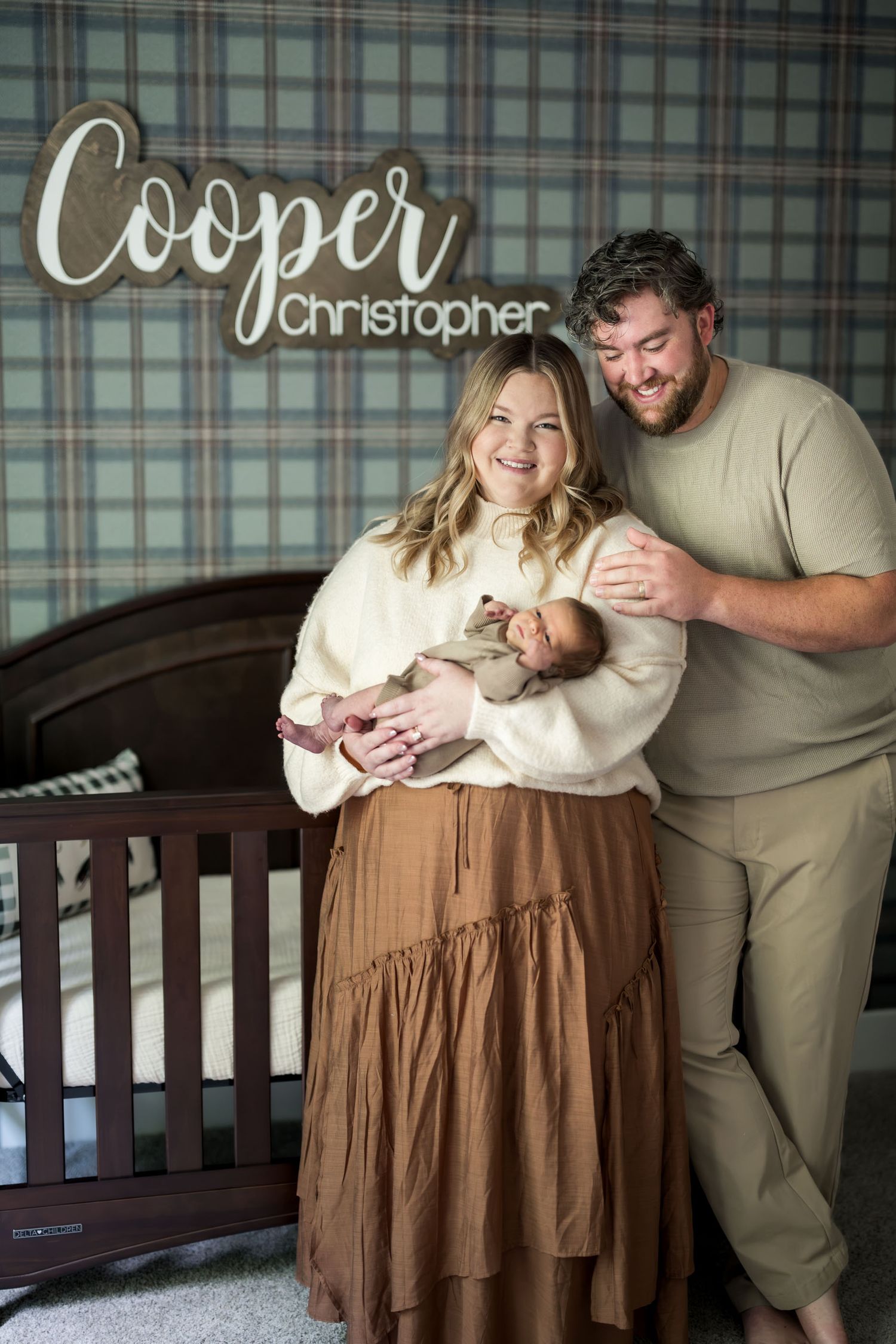 New parents smile proudly holding newborn baby Cooper Christopher in a plaid nursery.