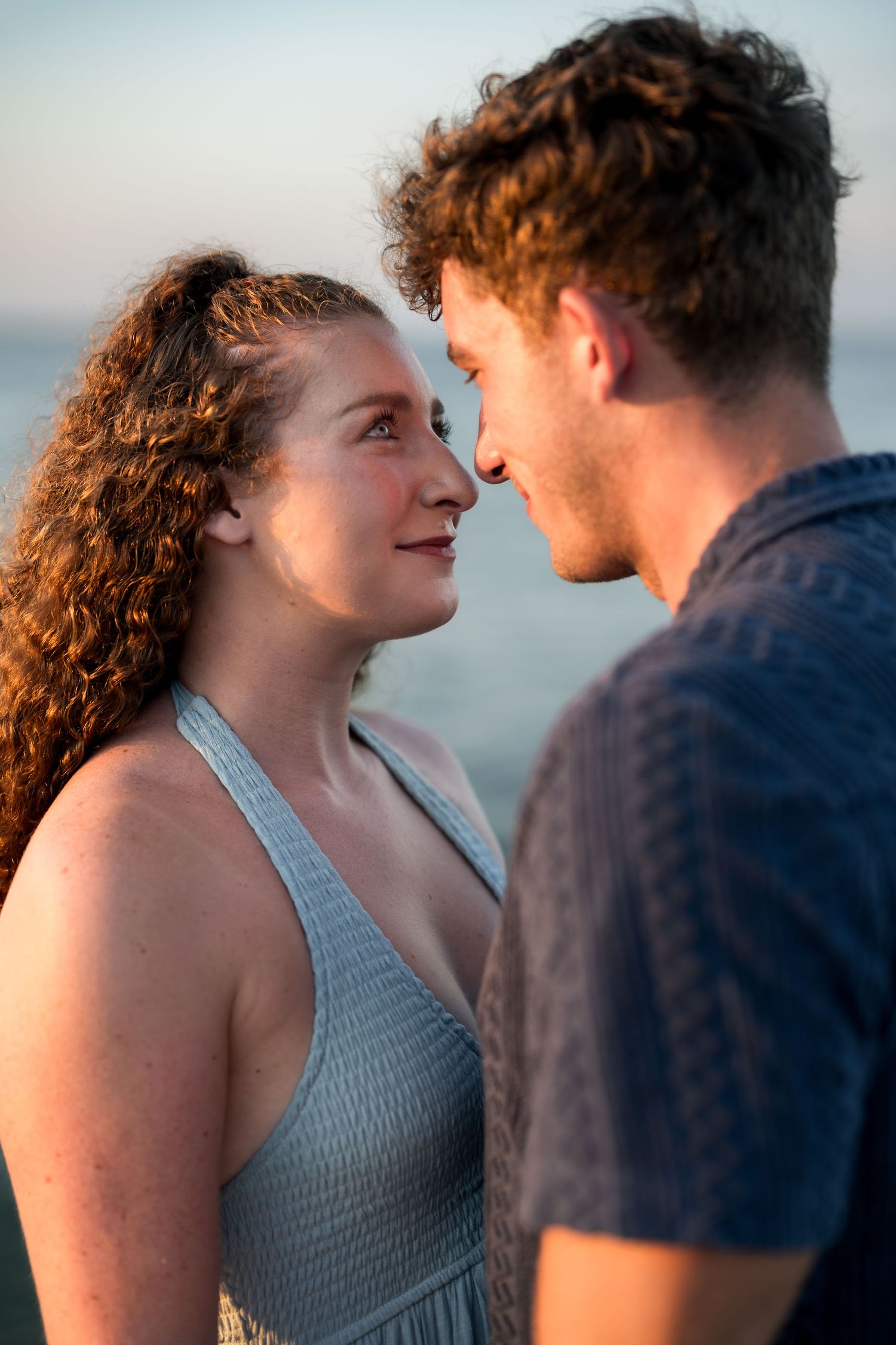 A couple shares a romantic moment gazing into each other's eyes near the ocean at golden hour sunset.