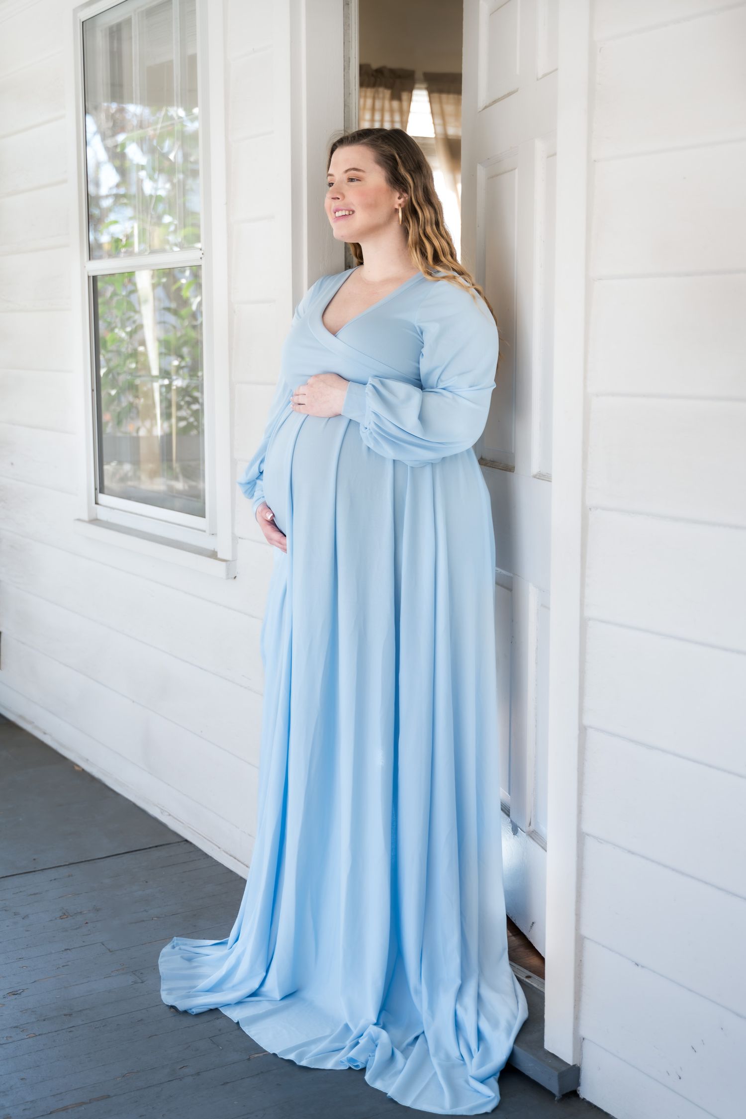 Smiling pregnant woman in elegant light blue long-sleeve maternity gown poses on a white porch holding her belly.