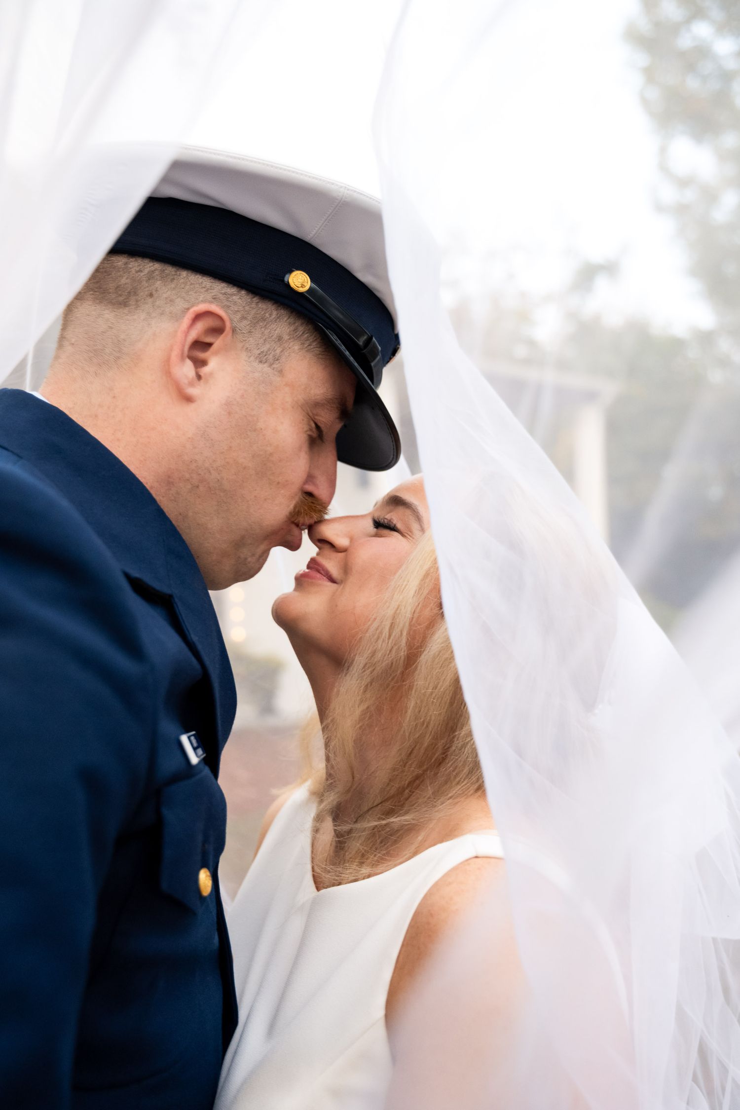 Romantic close-up of bride and groom almost kissing beneath flowing white wedding veil outdoors.