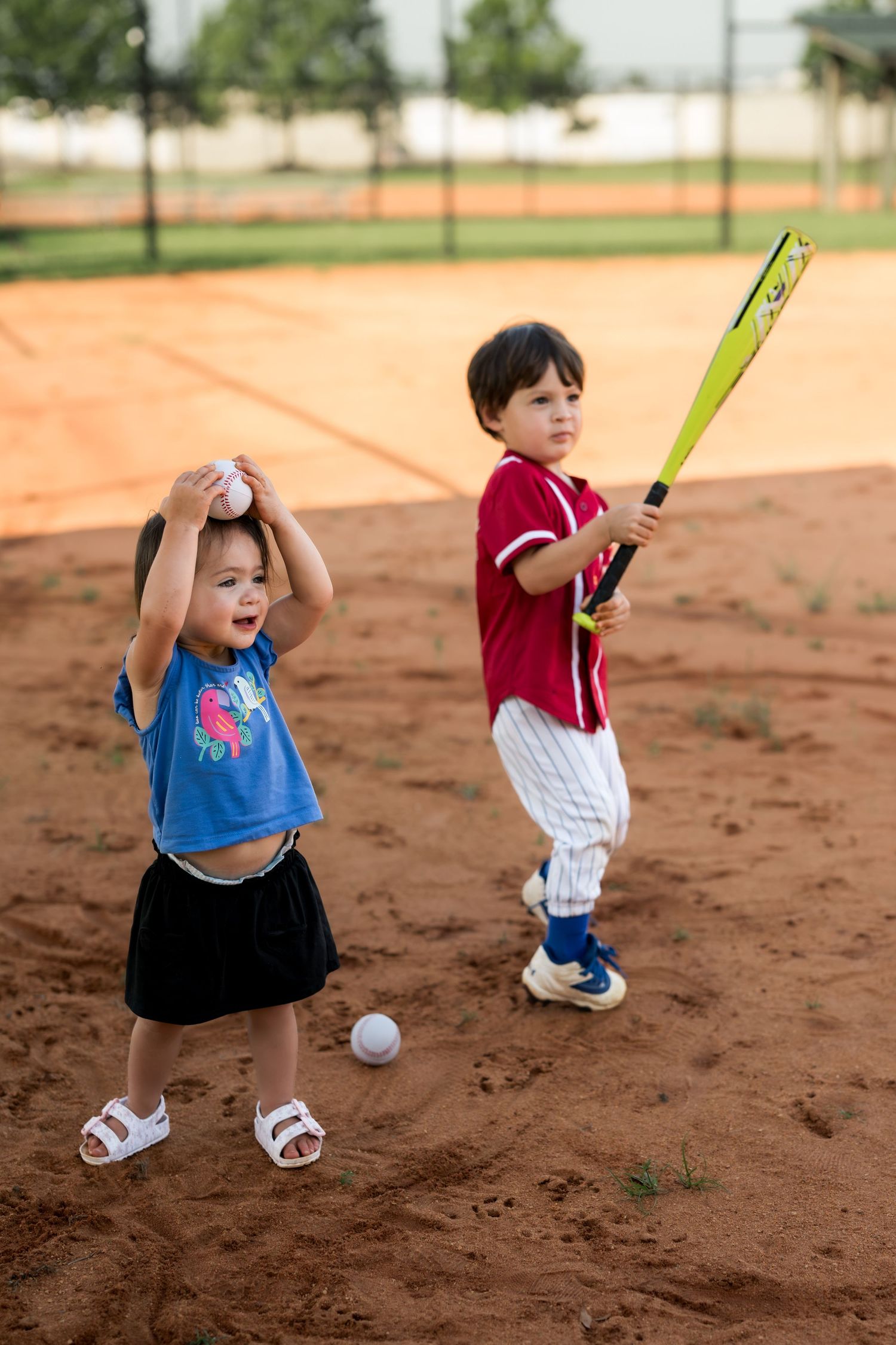 Two young children playing baseball on a dirt field, girl holding ball and boy in red uniform swinging yellow bat.