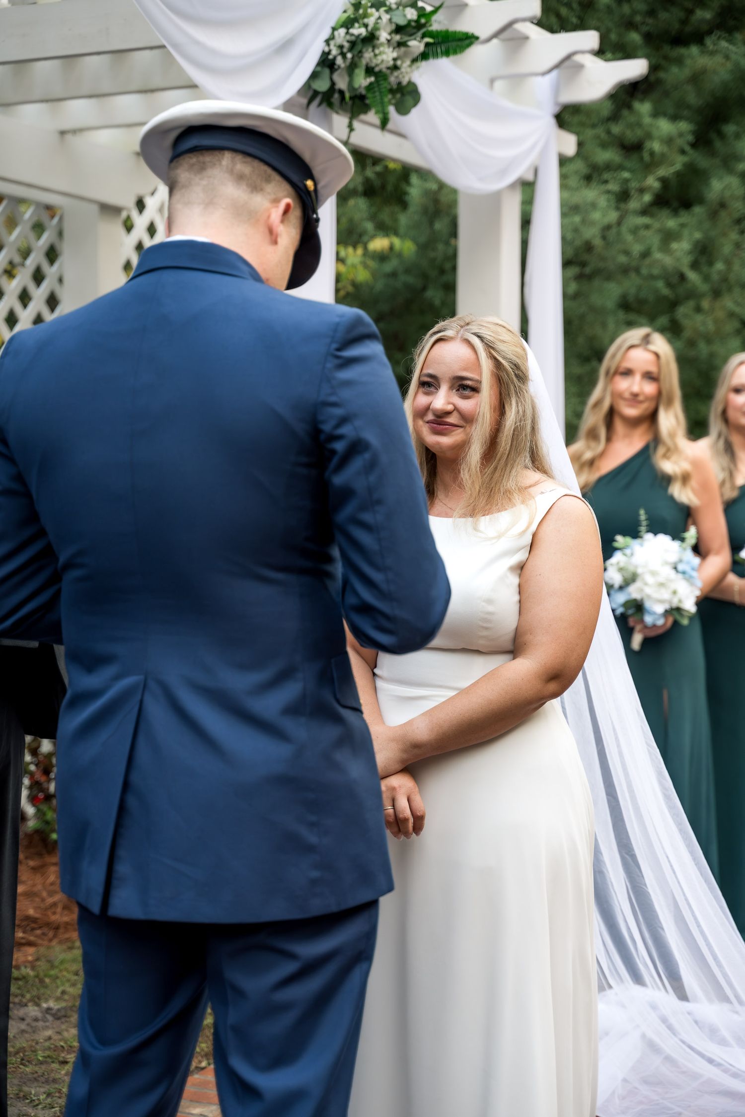 Bride in white gown holding hands with Coast Guard groom in blue uniform during outdoor ceremony.