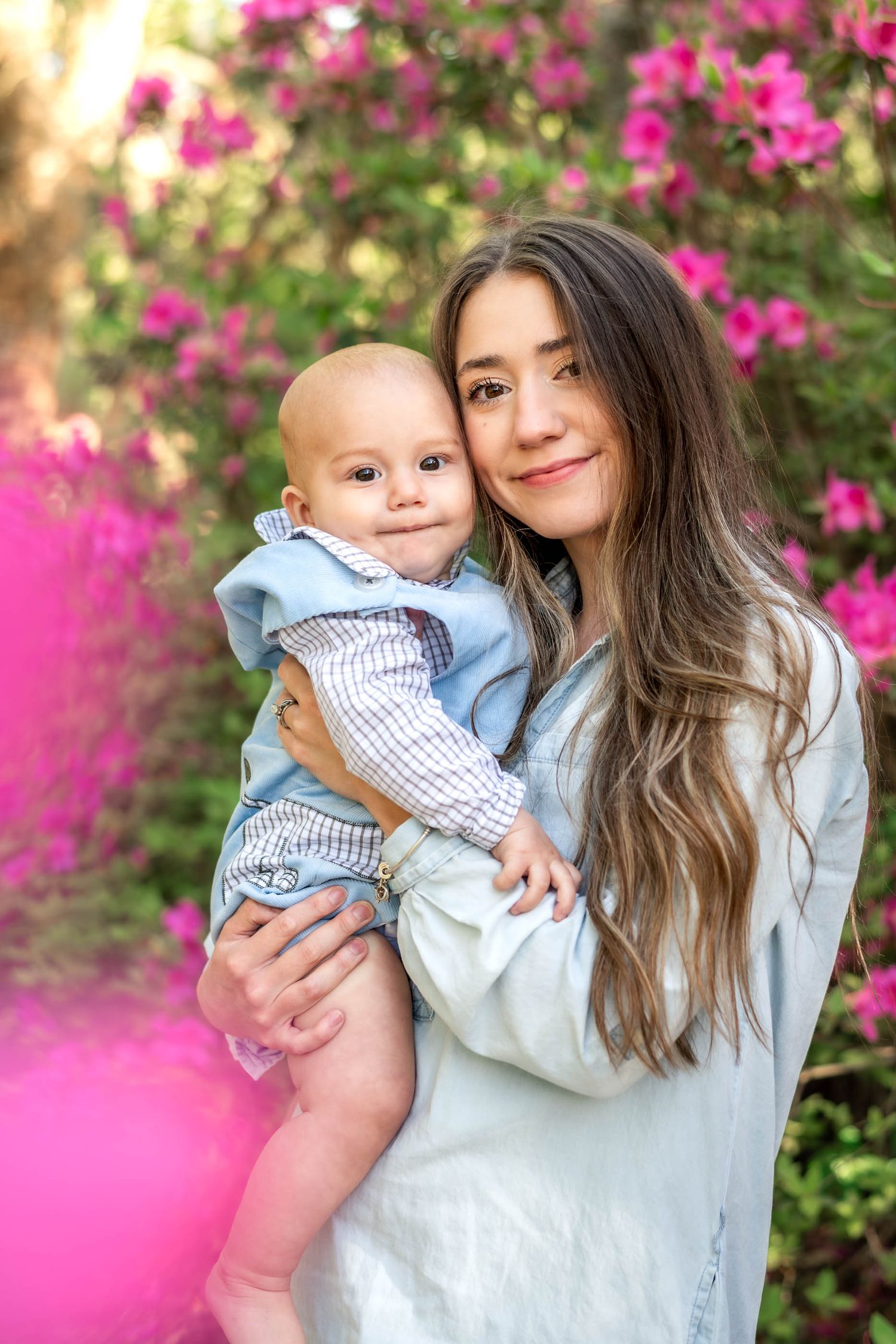 Young mother holding smiling baby boy in front of vibrant pink flowering bushes outdoors.