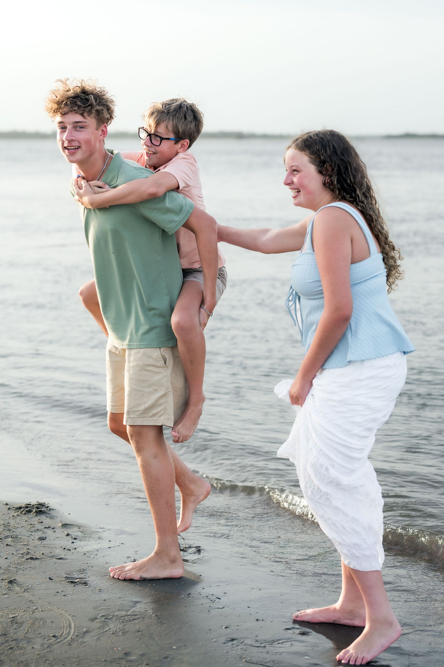 Three young people laughing and playing on a beach, with a boy giving a piggyback ride near the ocean waves.