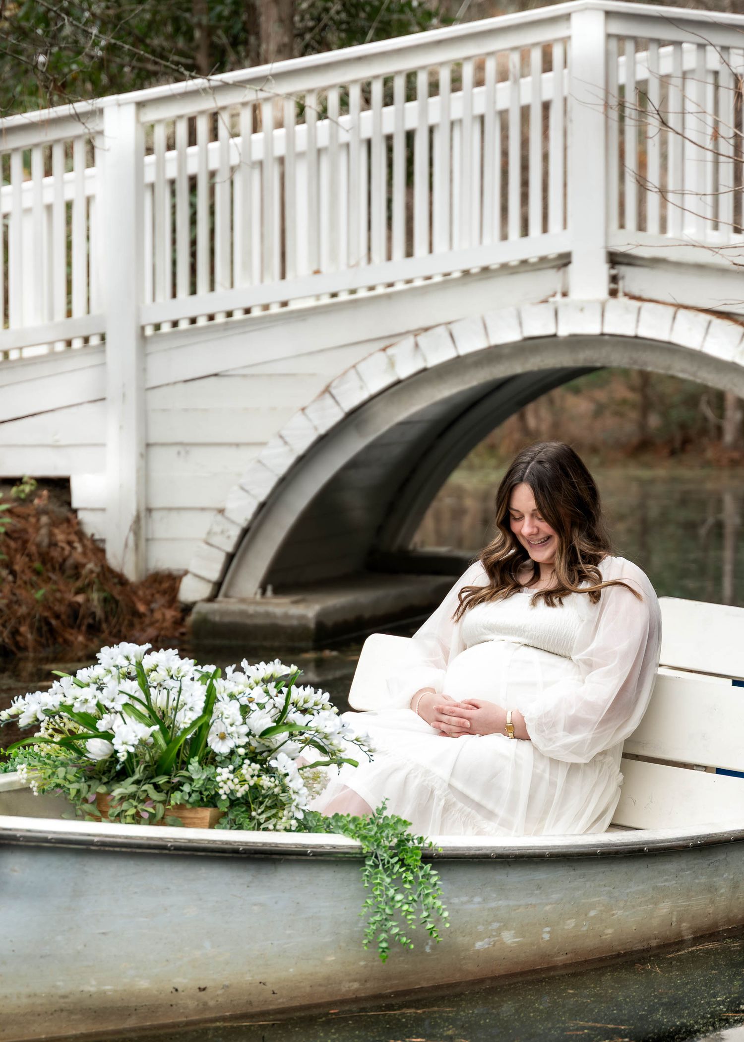 Cypress Gardens. Pregnant woman in white dress sits in a canoe with white flowers beside a charming white arched bridge outdoors.