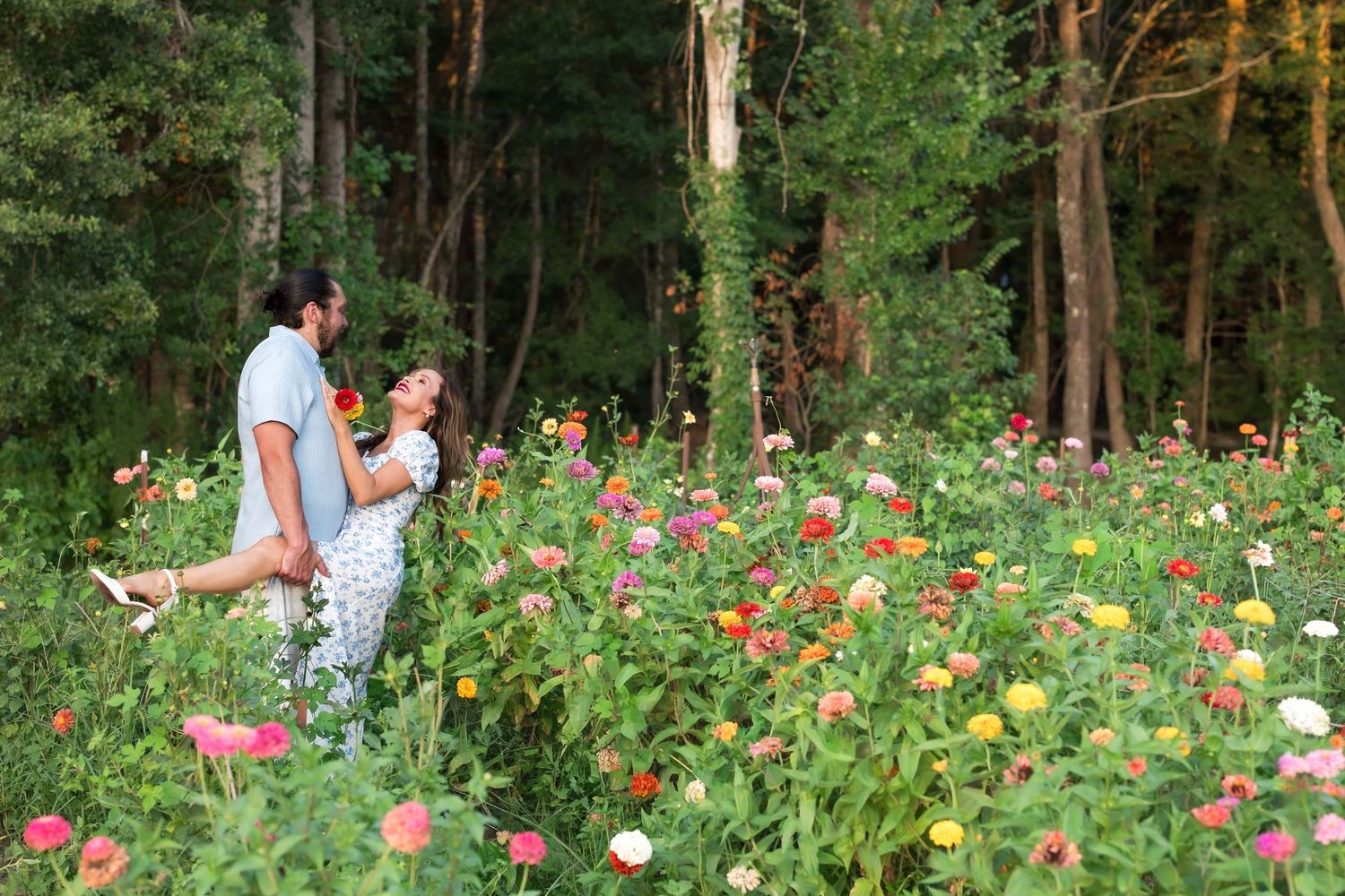 Couple embracing in a vibrant flower field with colorful zinnias, surrounded by lush green forest trees.