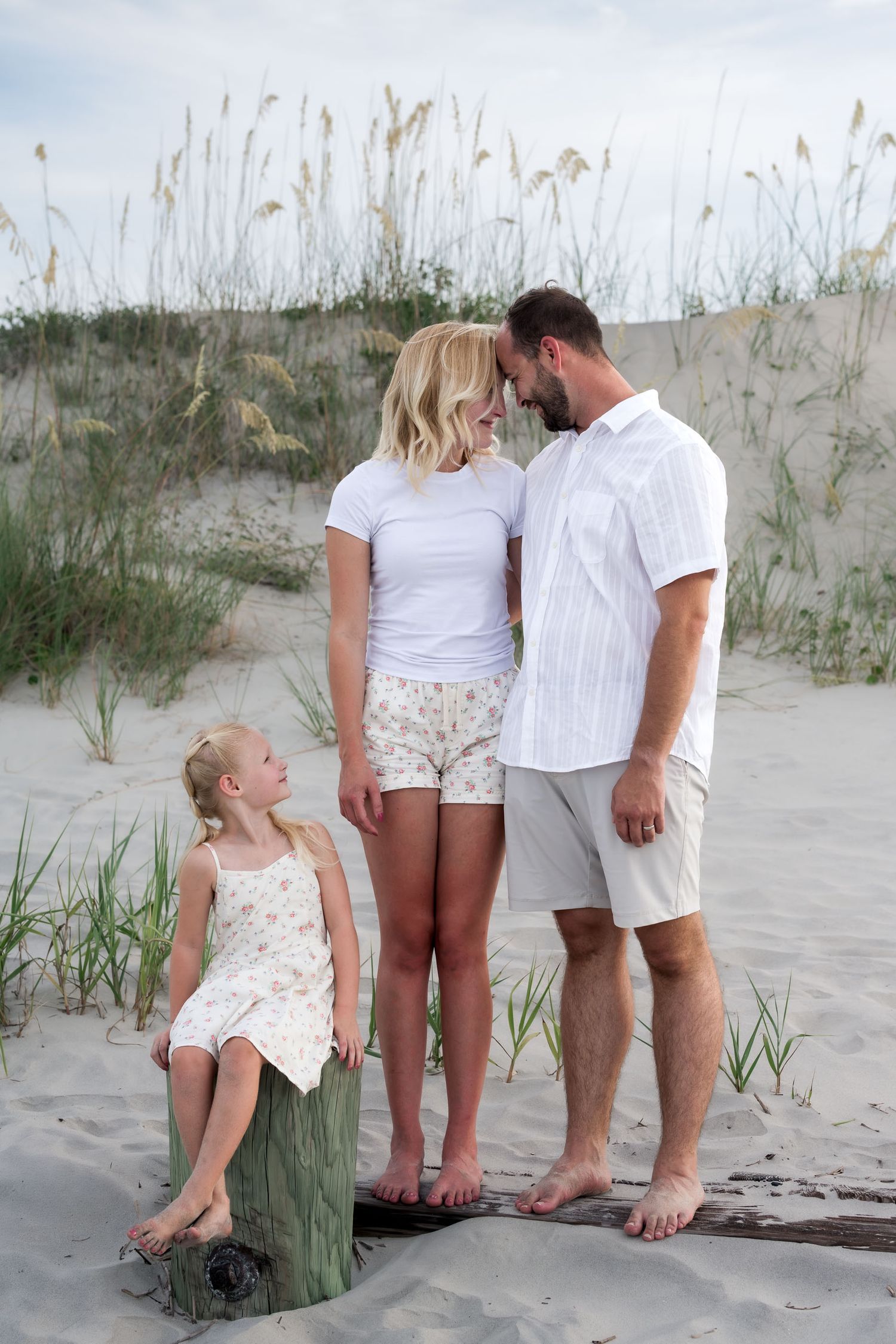 Family of three poses on sandy beach dunes, parents sharing a tender moment while young blonde girl sits on driftwood.