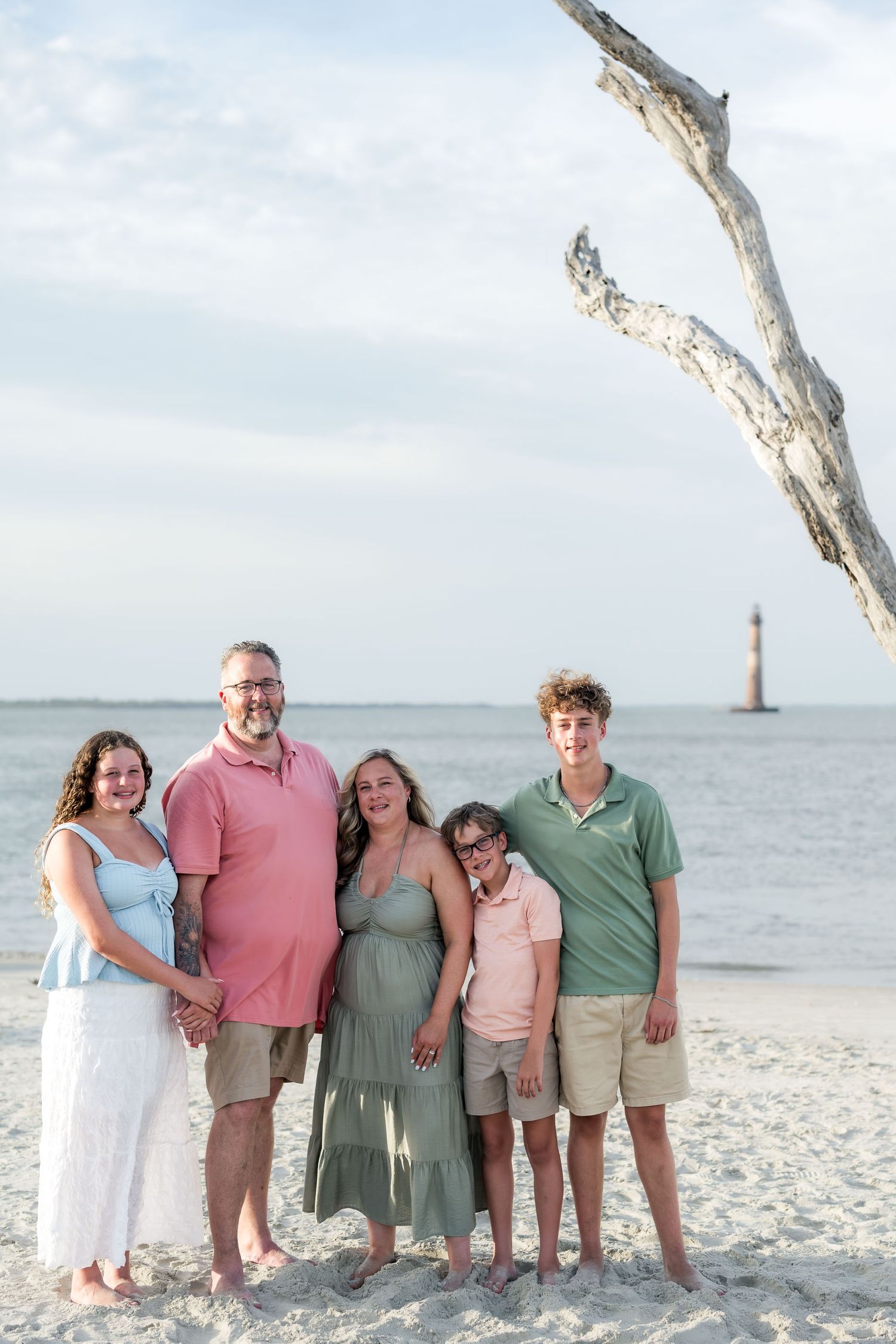Family of five posing on sandy beach near driftwood tree with lighthouse in background on a sunny day.