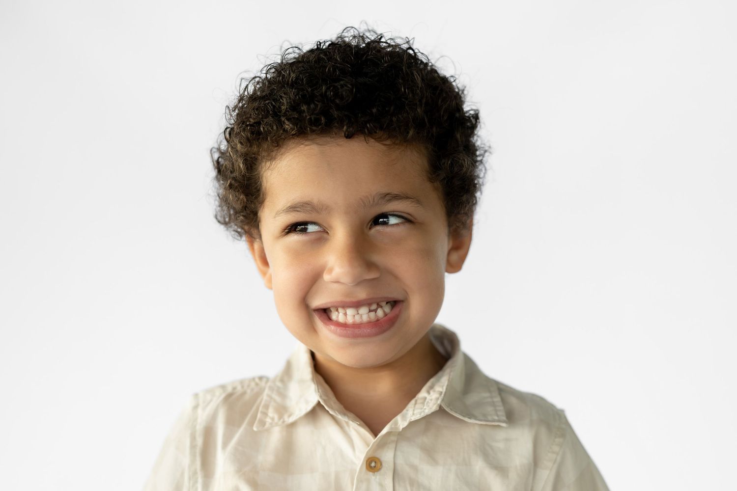 Smiling young boy with curly hair wearing a beige shirt against a white background.