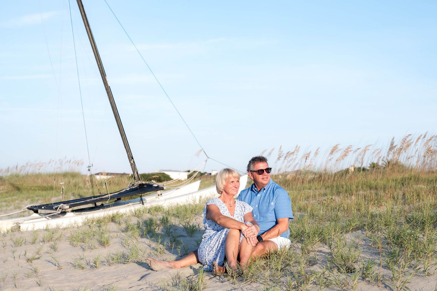Couple sitting together on sandy beach dunes near a sailboat, enjoying a sunny day with sea oats in the background.
