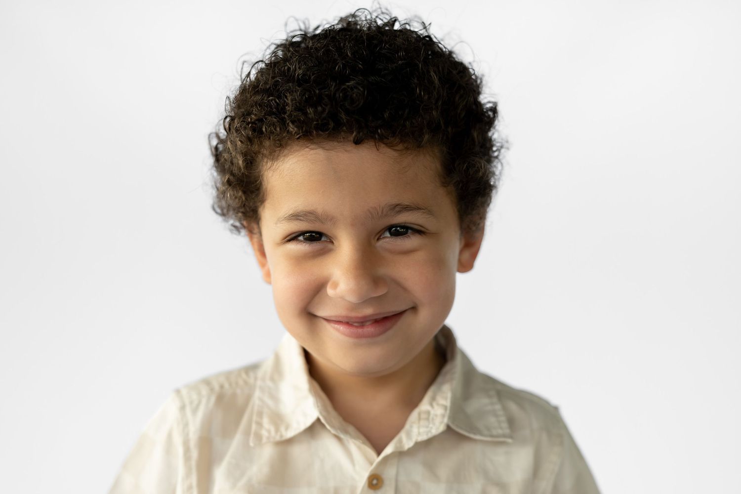 A young boy with curly hair giving a gentle smile, wearing a light beige shirt against a white background.