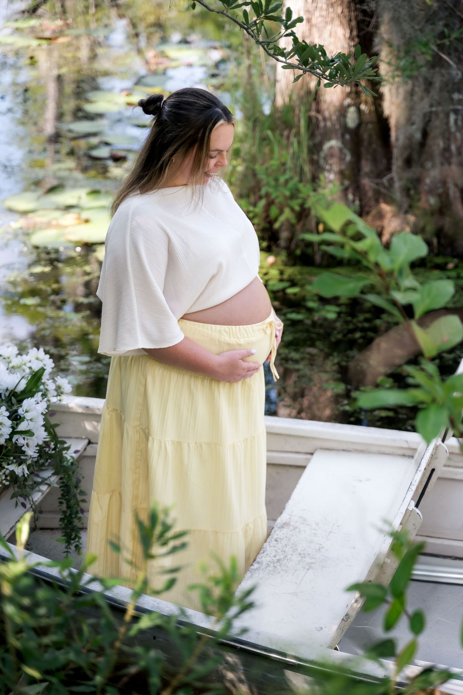 Pregnant woman in white crop top and yellow skirt cradling baby bump outdoors near a lily pond.