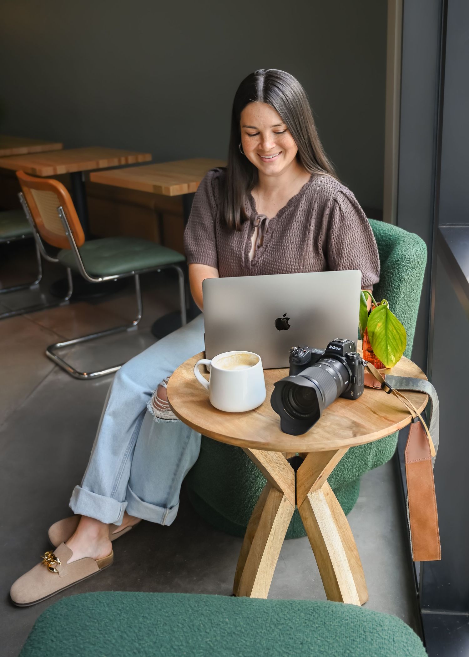 A smiling woman works on a MacBook laptop beside a professional camera and coffee in a cafe.