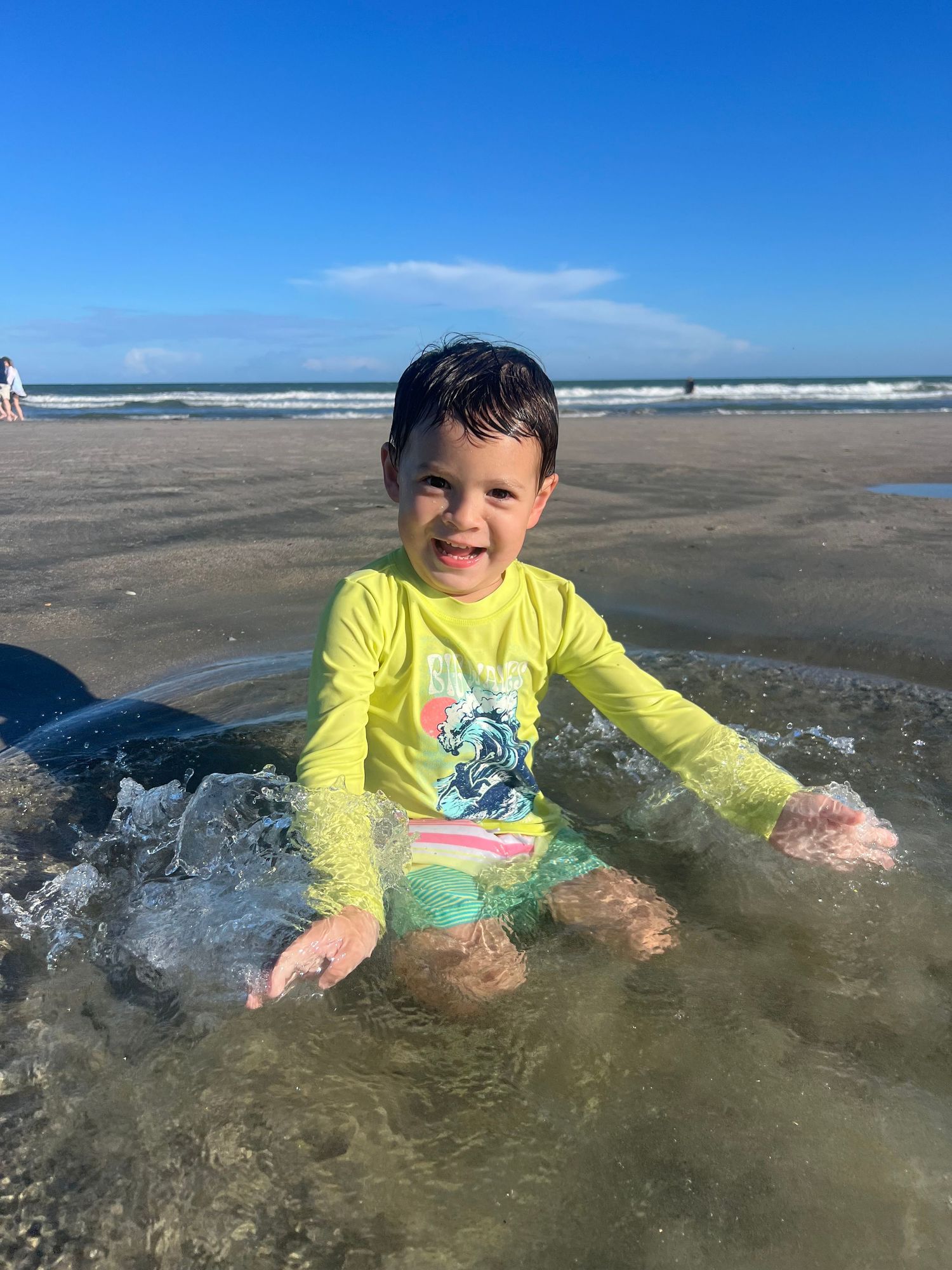 Happy young child in yellow rash guard splashing in ocean waves on a sunny beach day.