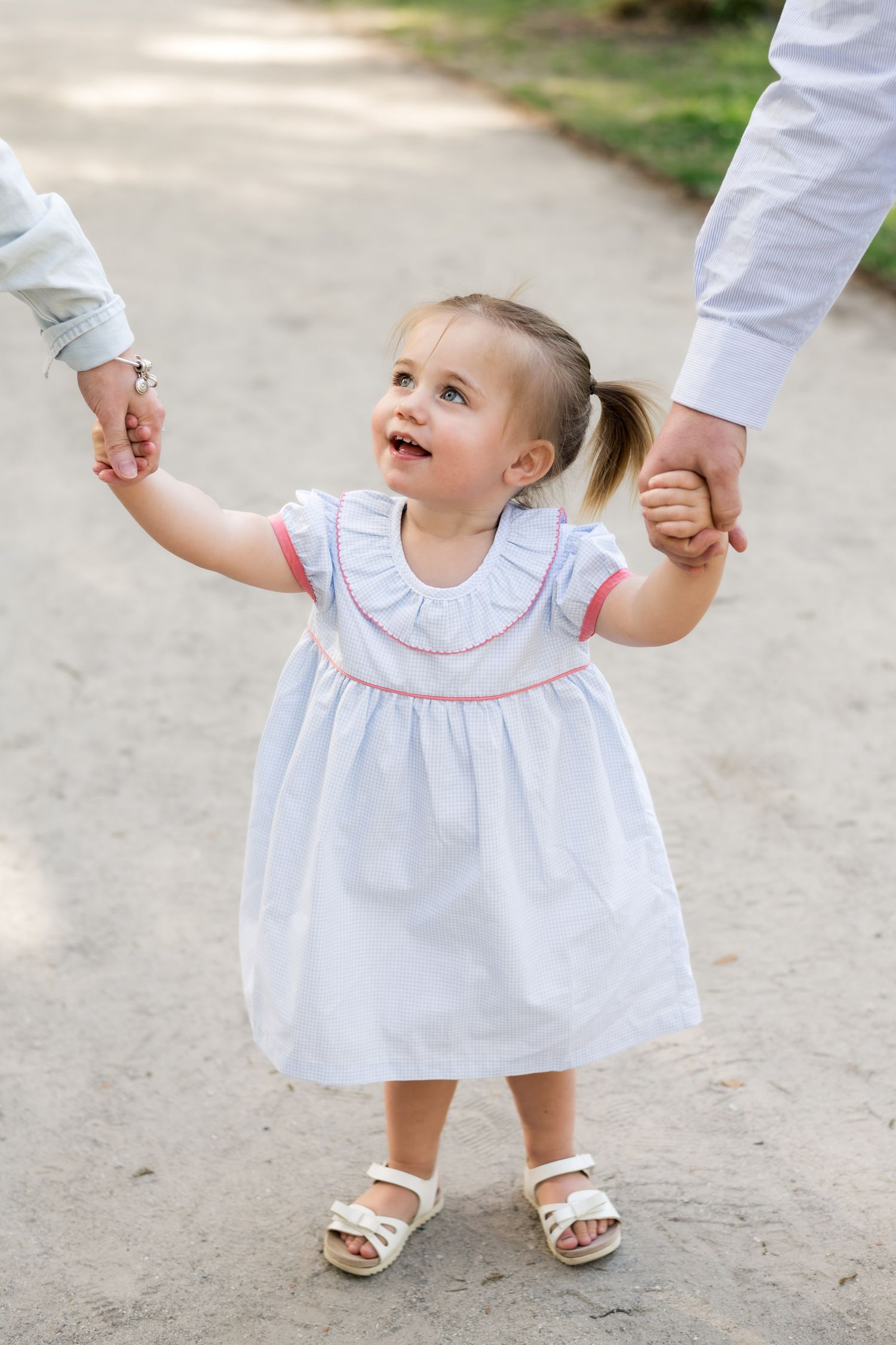 A toddler girl in a white dress with pink trim holds hands with two adults while walking outdoors.