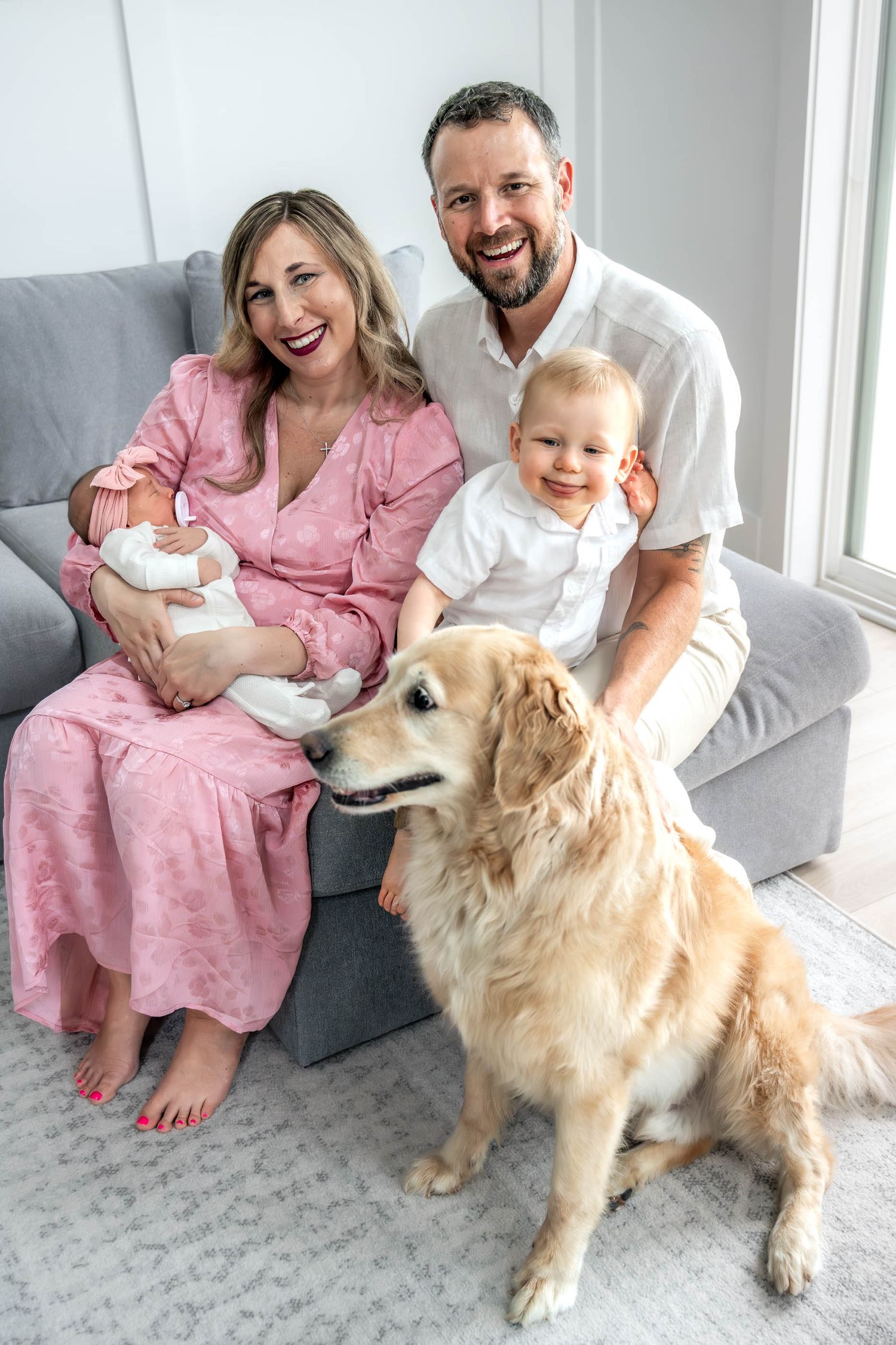 Happy family portrait with parents, toddler, newborn baby, and golden retriever dog on gray sofa.