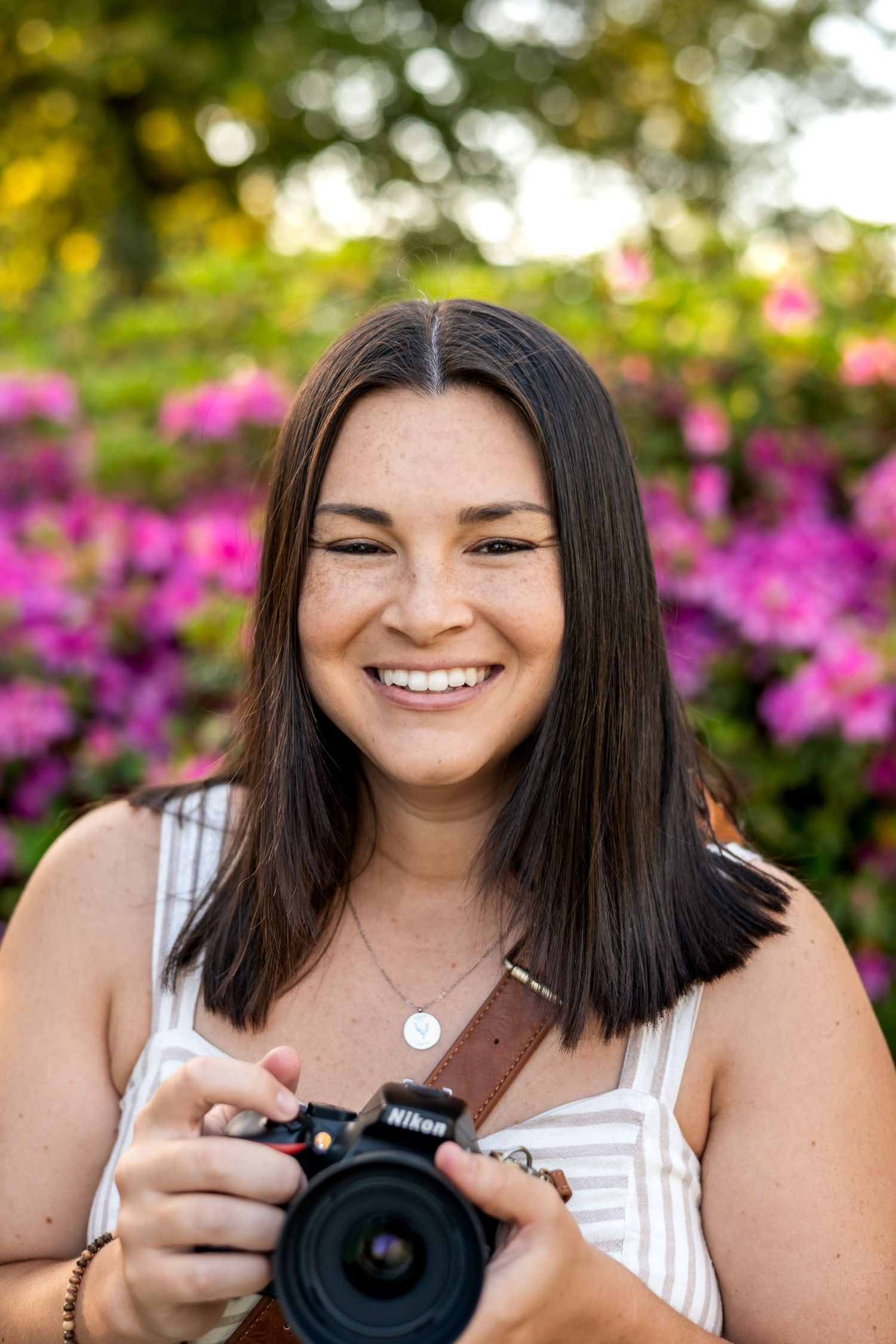Smiling female photographer with freckles holding a Nikon camera with pink flowers behind her.