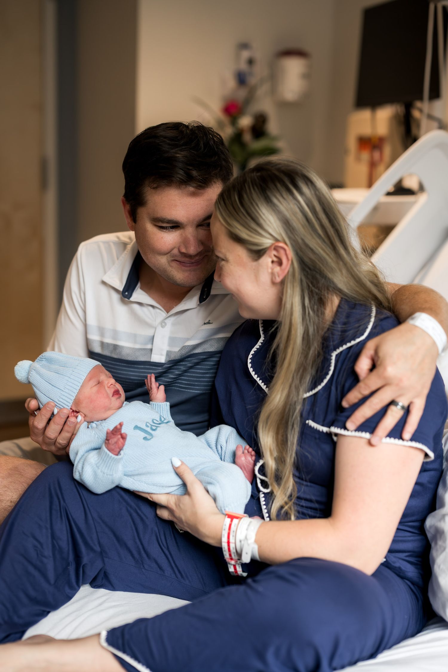 New parents smile lovingly at their newborn baby in a hospital room, mother in navy robe, father in striped shirt.